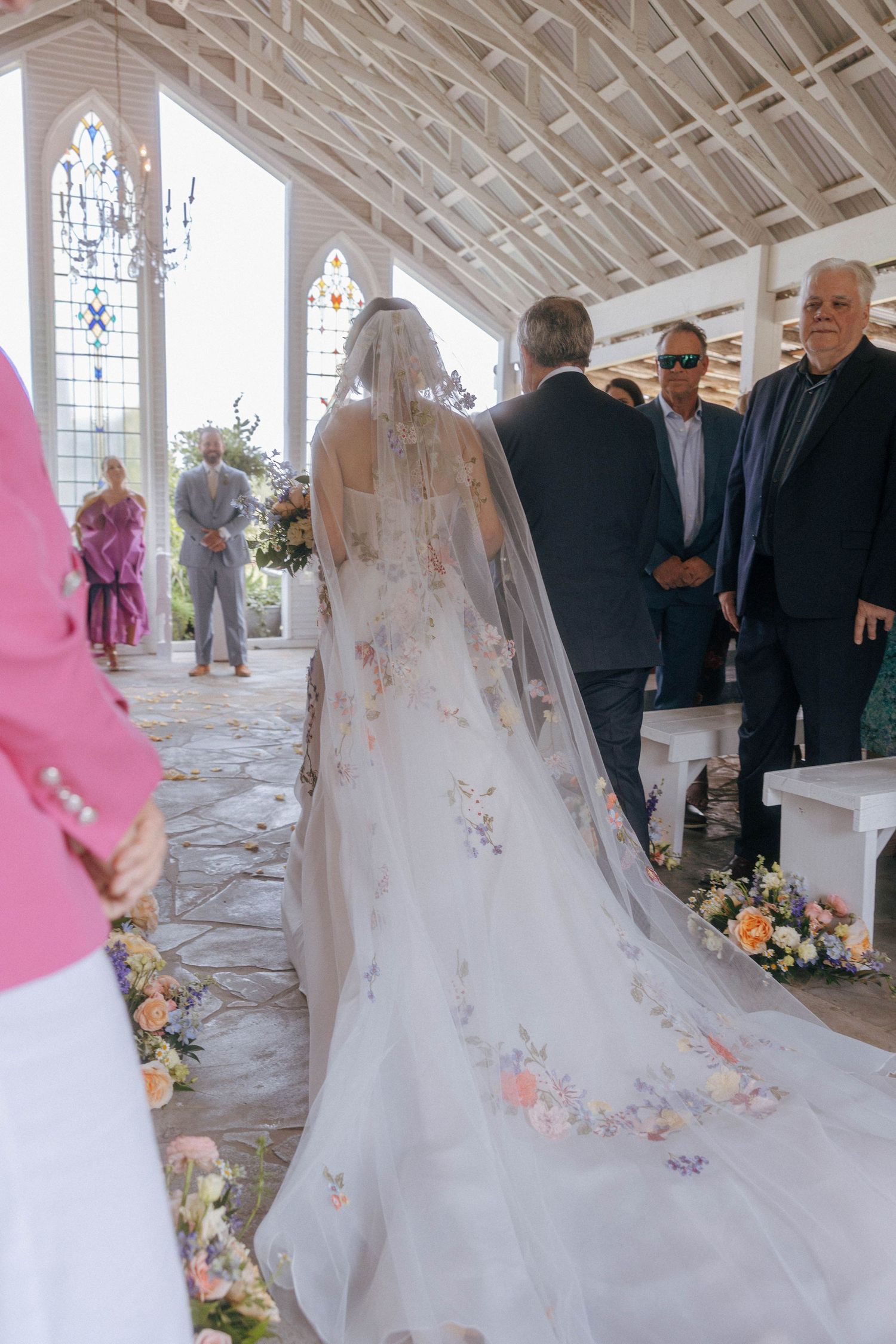 Bride walks down aisle in floral embroidered cathedral veil inside a white chapel.