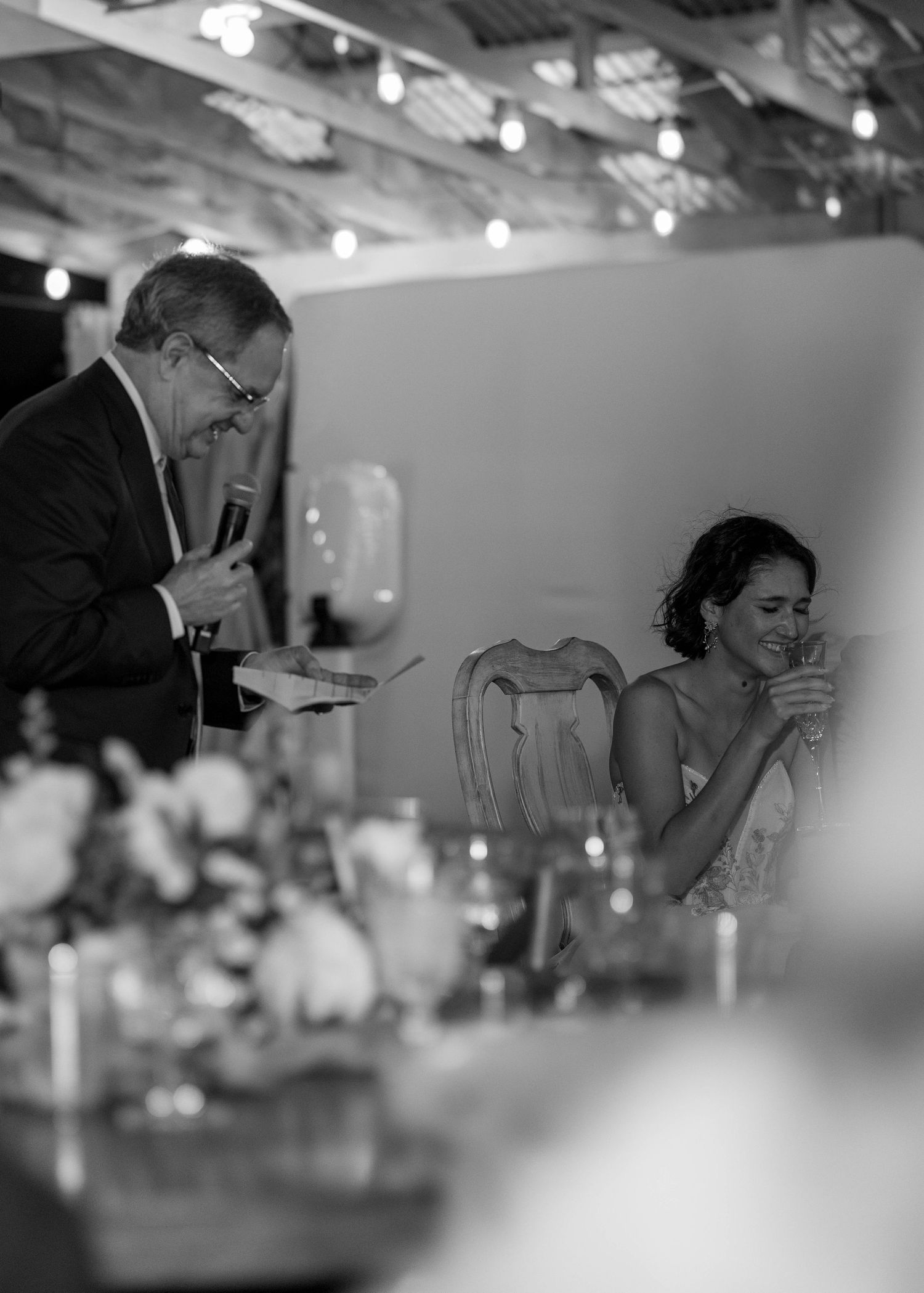 Black and white photo of father giving speech at wedding while bride laughs at head table.