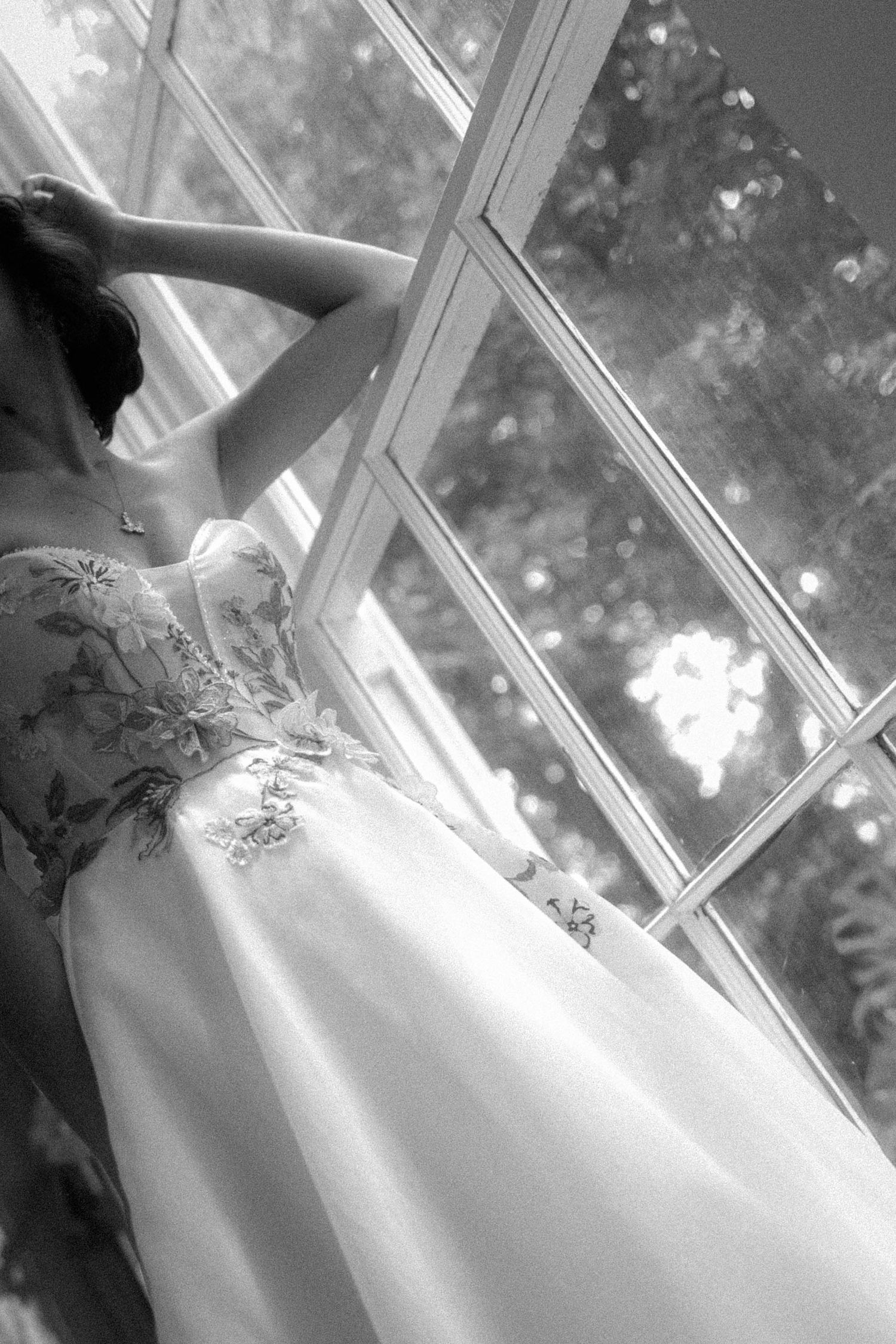 Black and white photo of a bride in a floral strapless gown reaching up near a sunlit window.