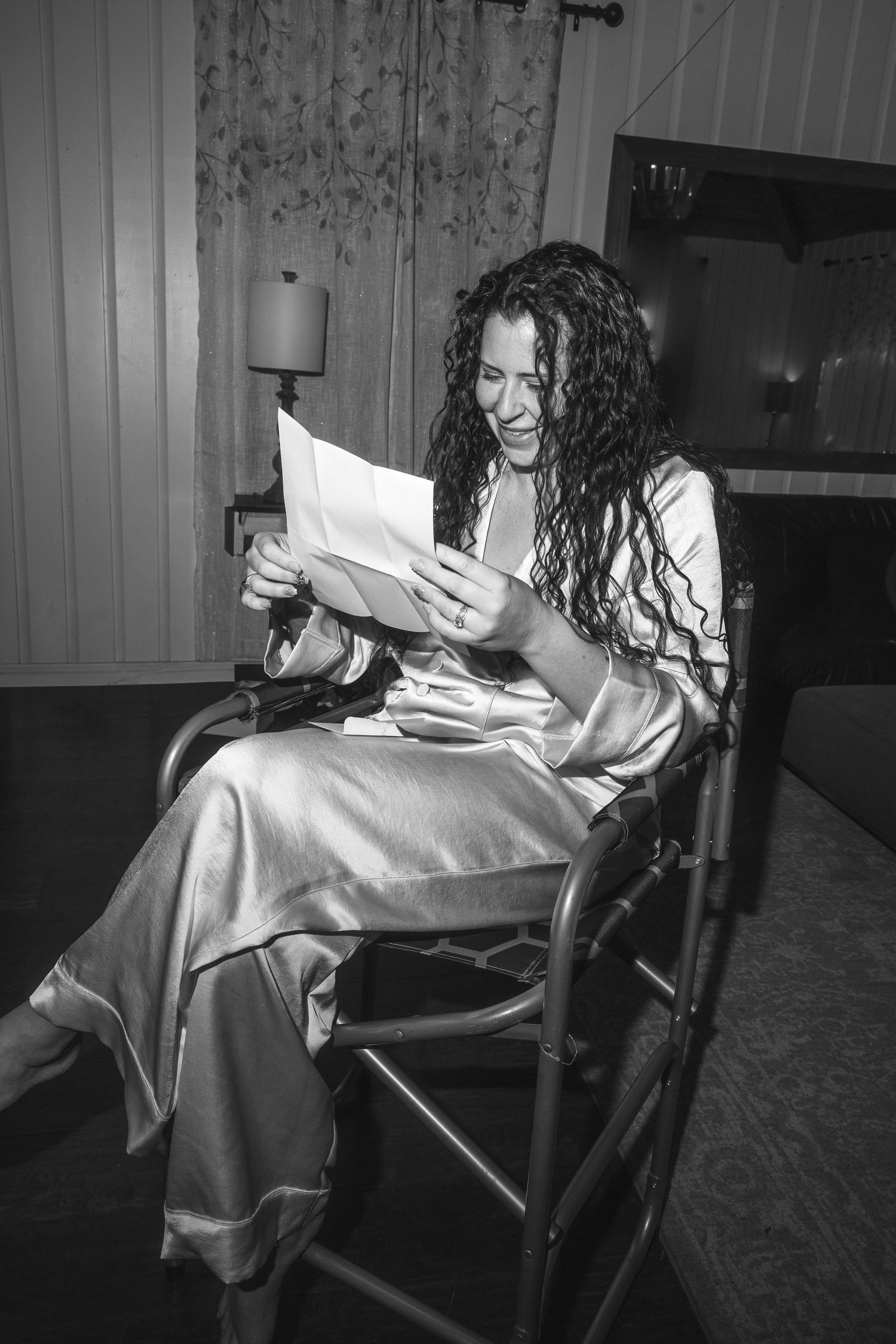 Black and white photo of curly-haired woman in satin robe smiling while reading a letter in a chair.