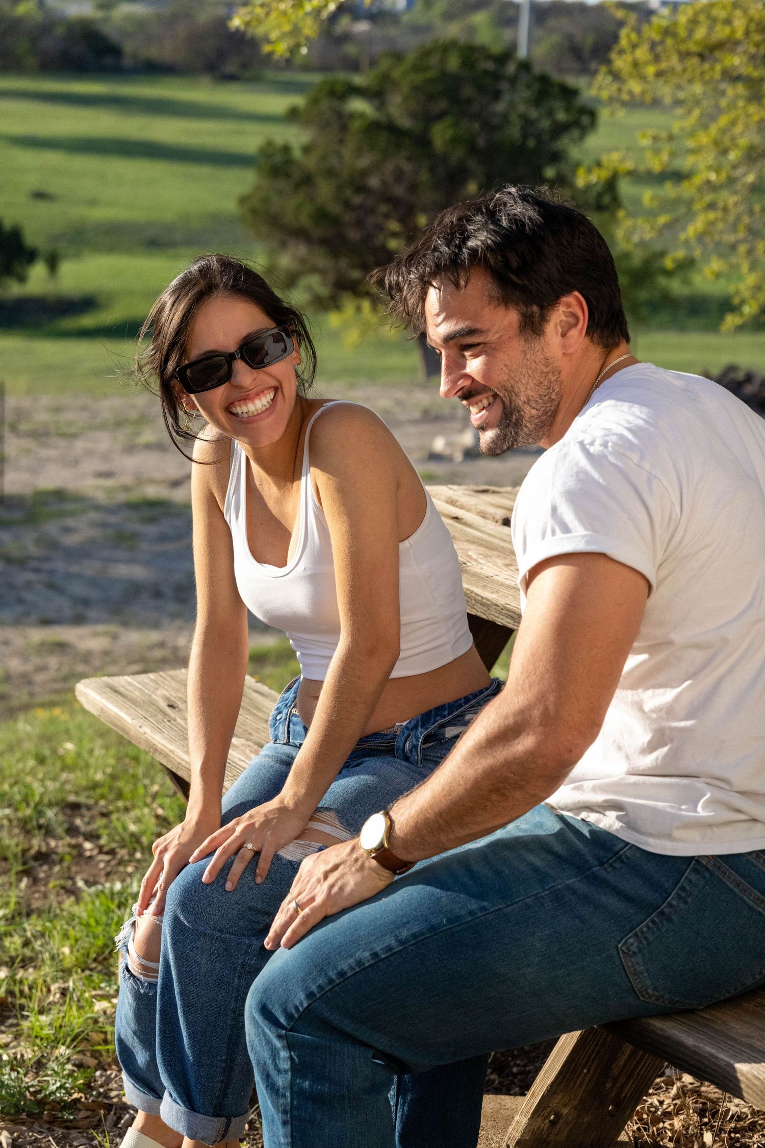 A smiling couple in casual white tops and jeans sit together on a wooden picnic table in a sunny green field.