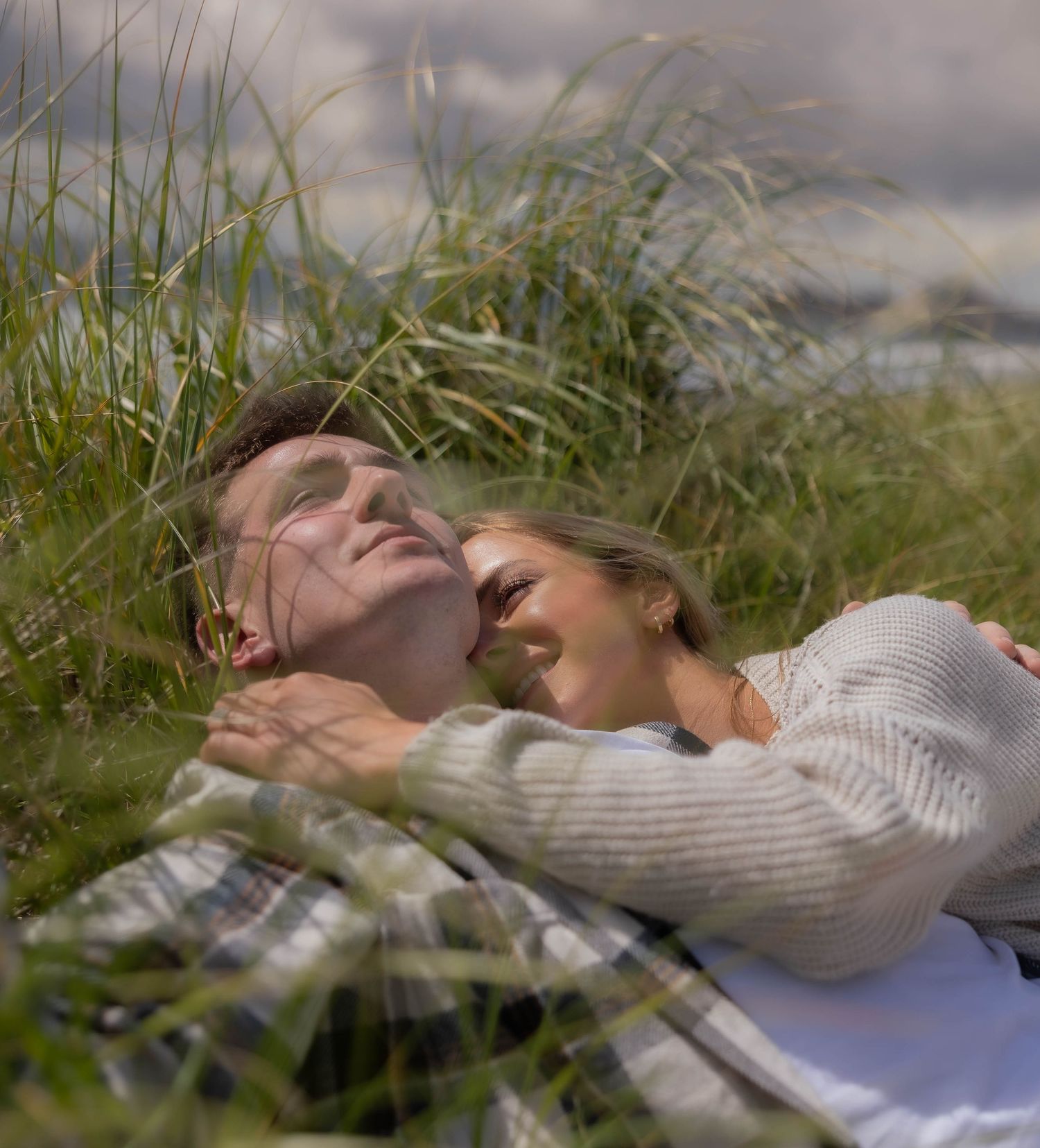 Couple lying together in tall green beach grass, woman smiling and embracing man in a peaceful moment.