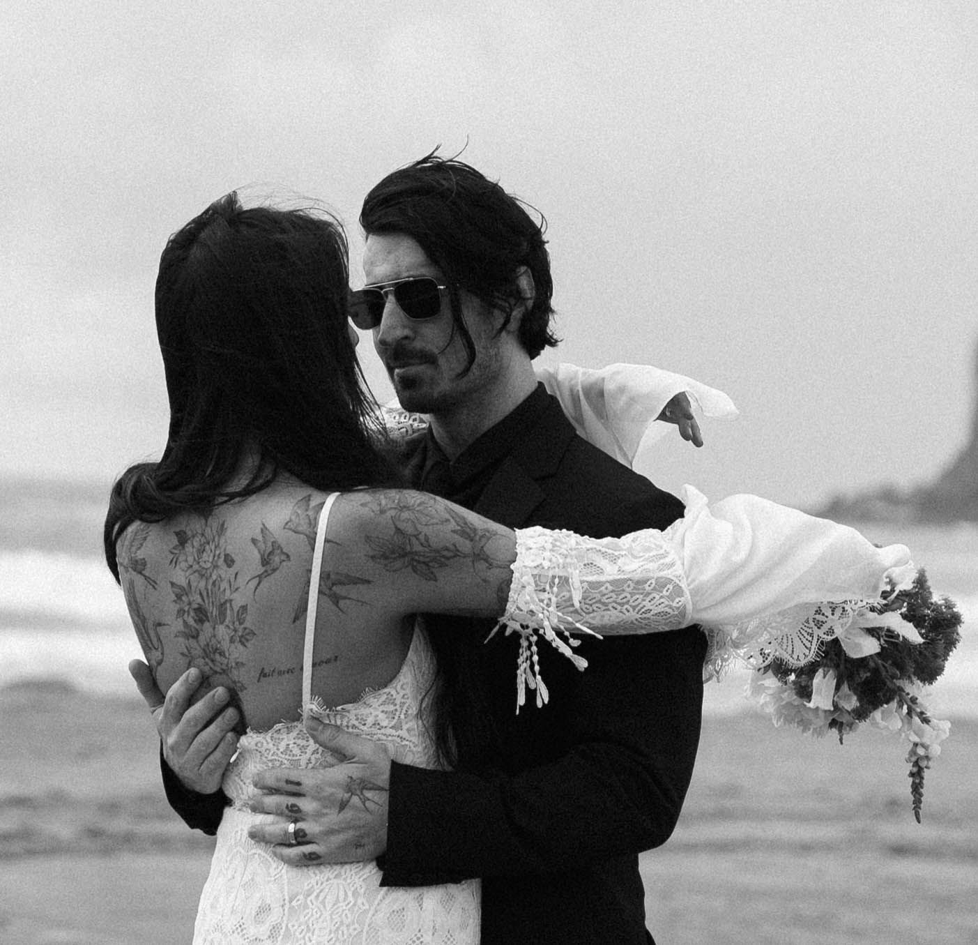 A couple in wedding attire embrace on a beach, with the bride in white lace and groom in dark suit and sunglasses.