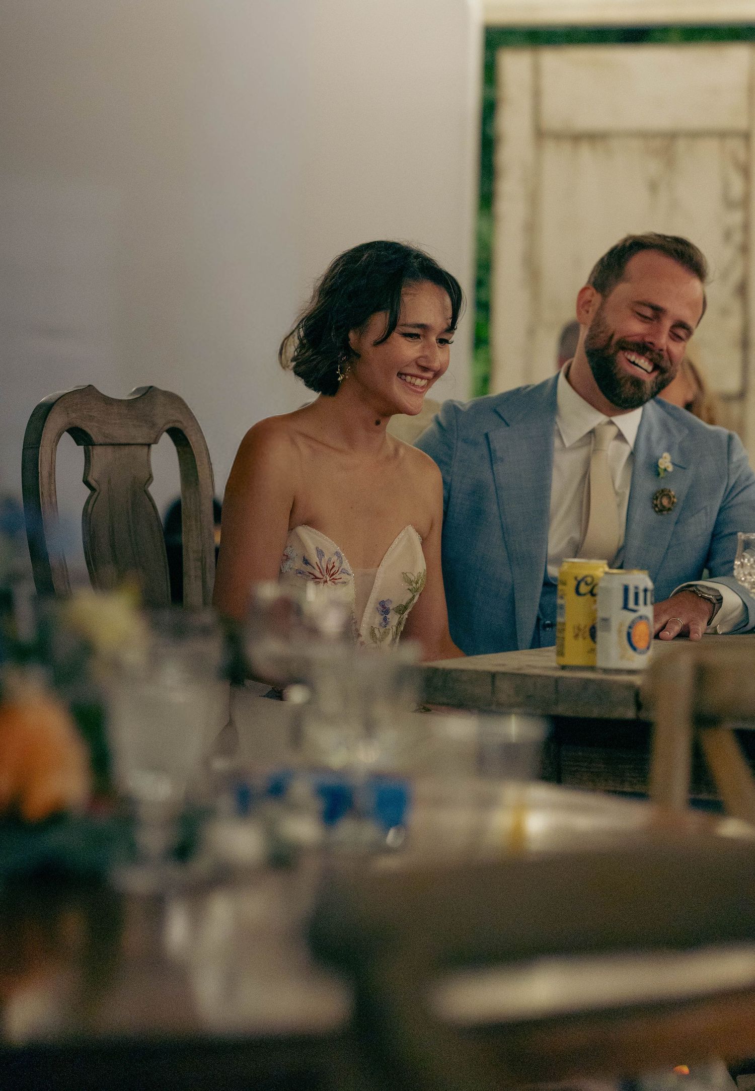 Bride and groom laughing together at wedding reception head table with Miller Lite cans nearby.