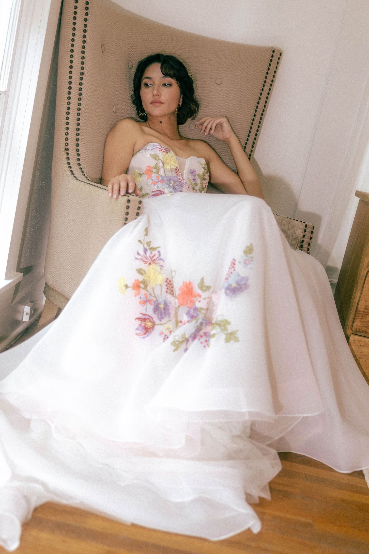 Bride in a white strapless ball gown with colorful floral embroidery seated in an elegant chair.