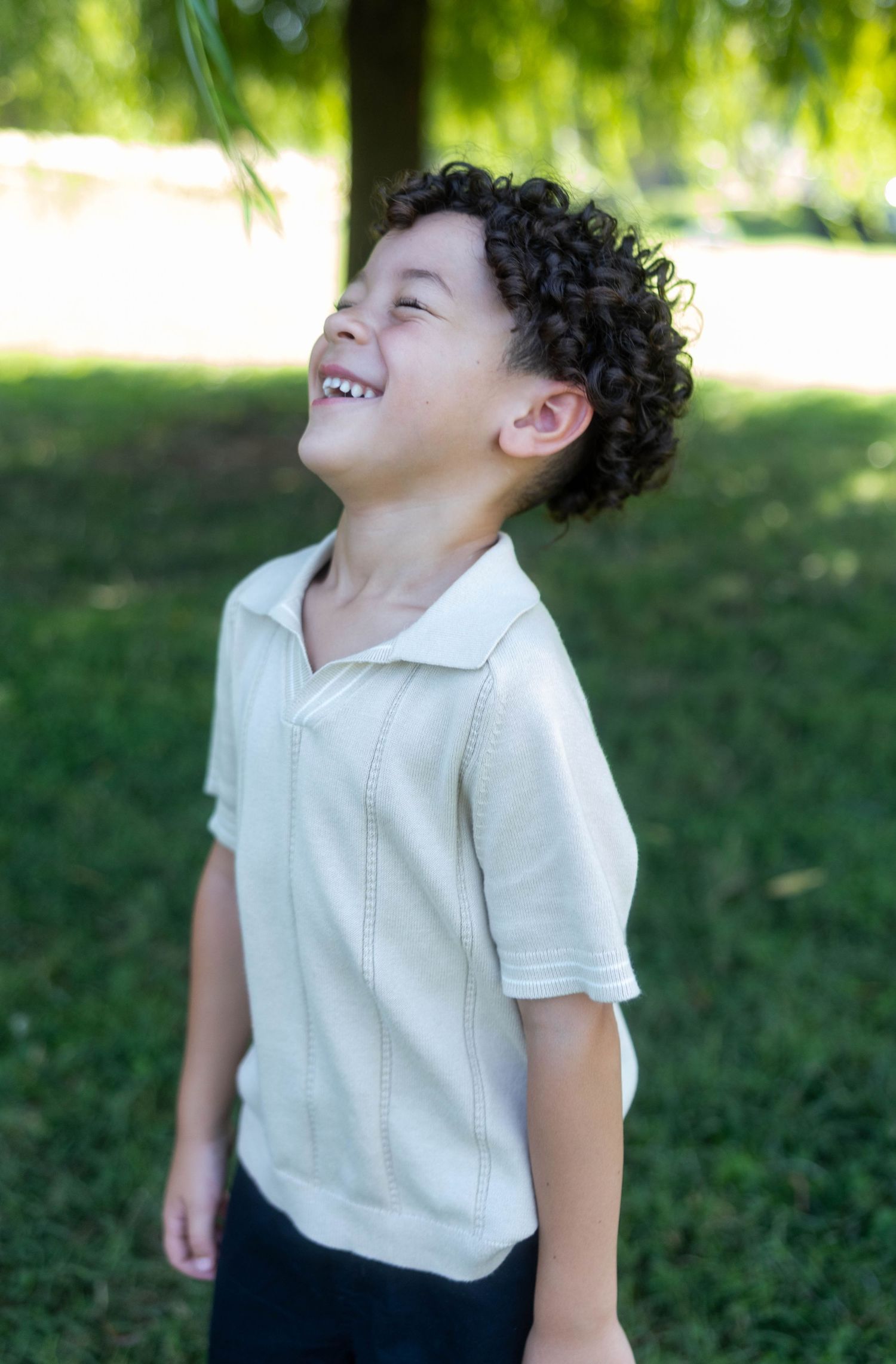 A joyful young boy with curly hair laughs outdoors, wearing a cream polo shirt against a green park background.