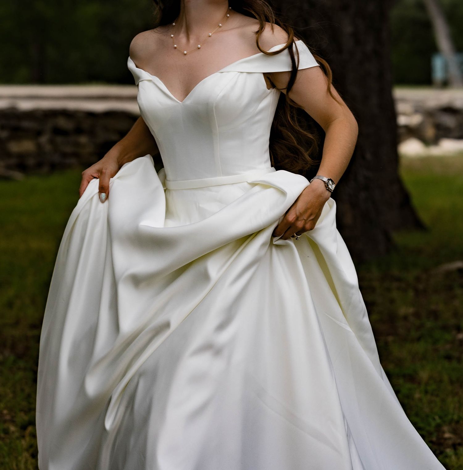 Bride in off-shoulder white ballgown holding skirt outdoors near a stone wall.