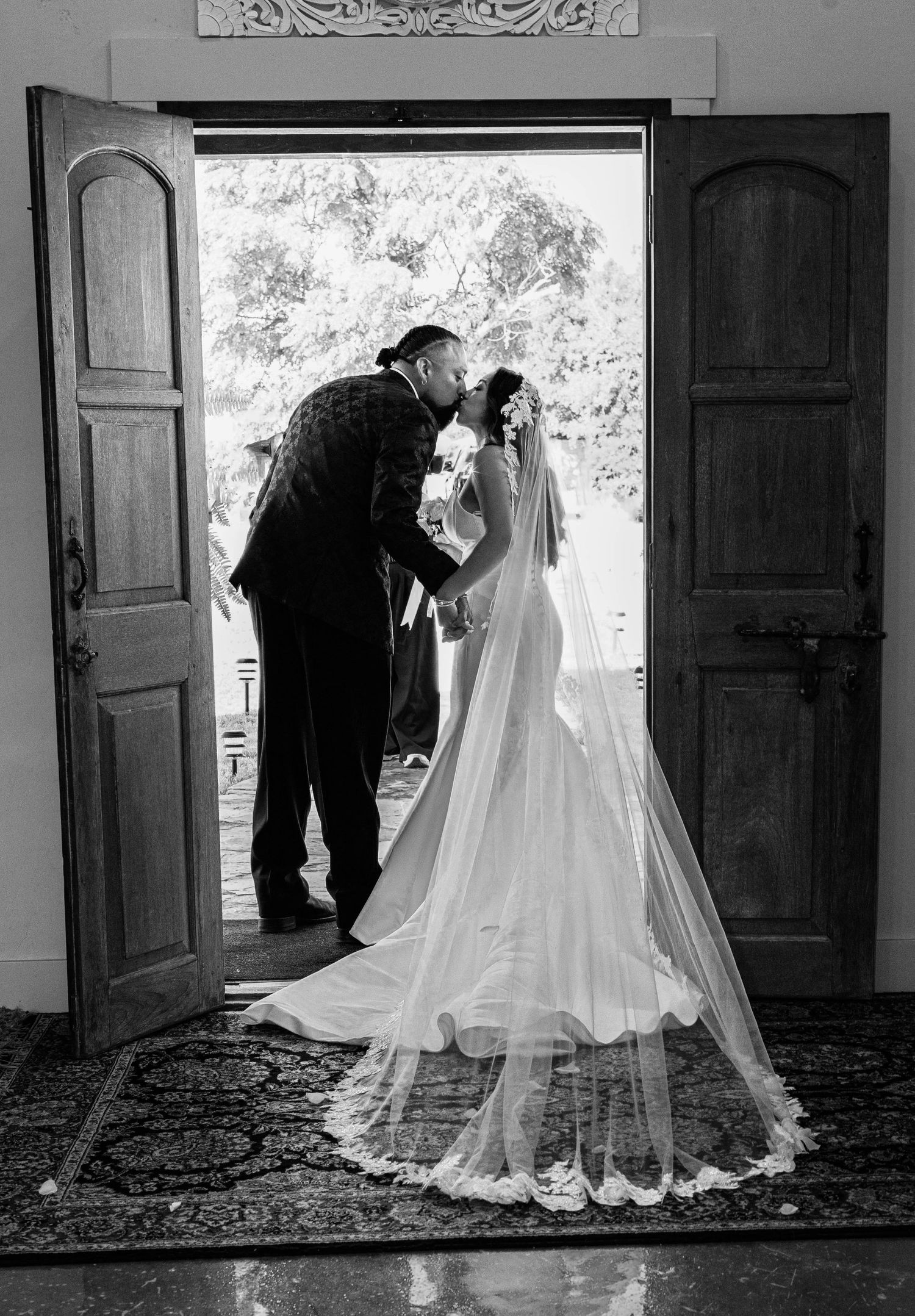 Black and white silhouette of groom kissing bride in cathedral veil at open church doors.