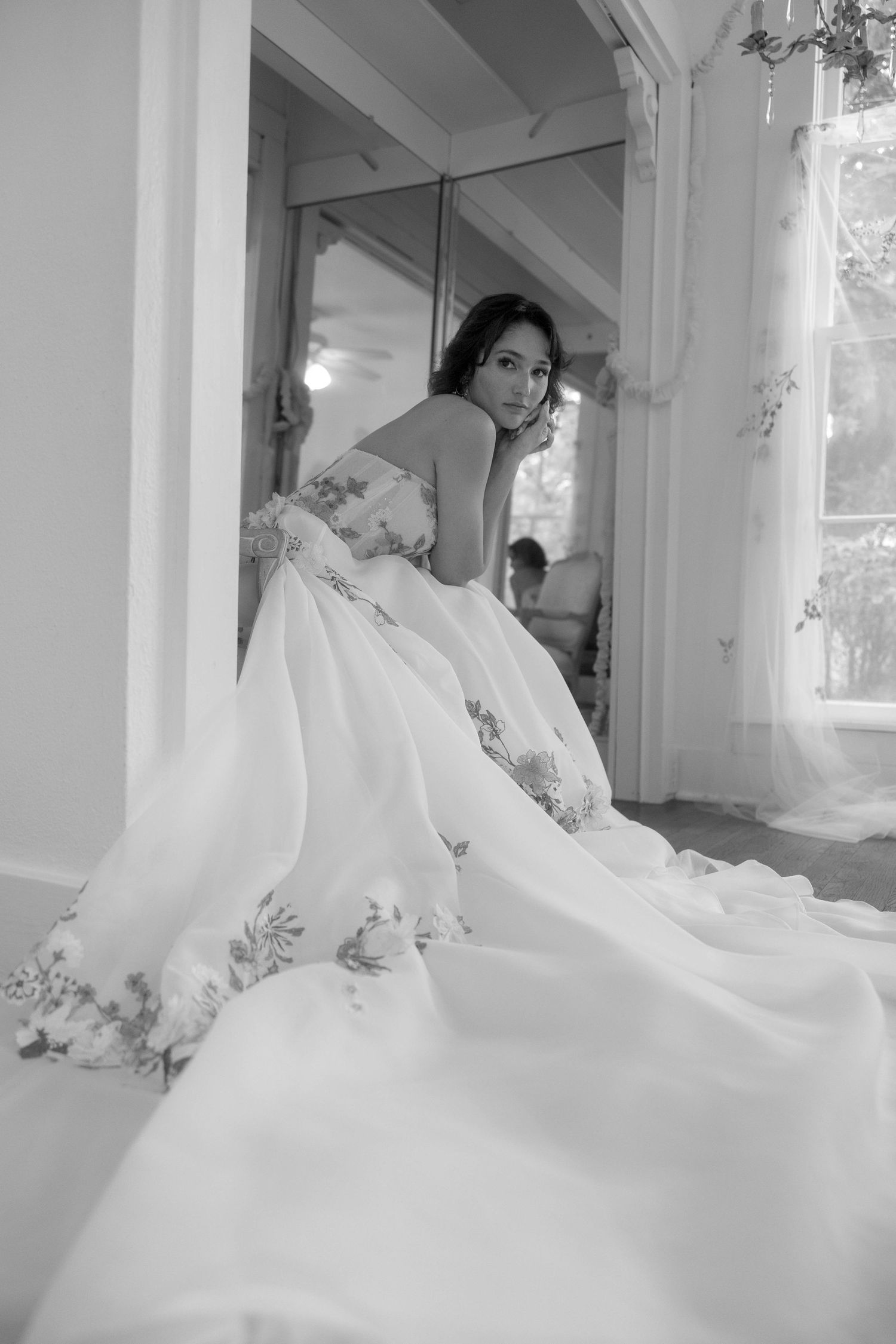 Black and white portrait of a bride in a floral ball gown seated elegantly by a mirror.