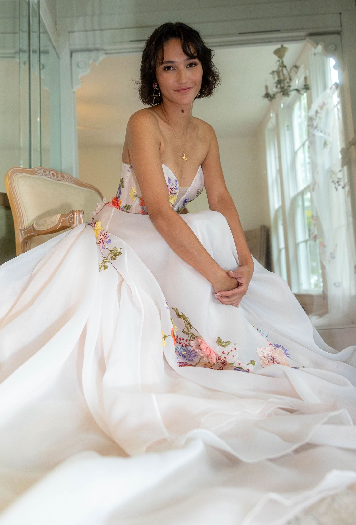 A smiling bride in a floral embroidered strapless ball gown sits gracefully in an elegant bridal suite.