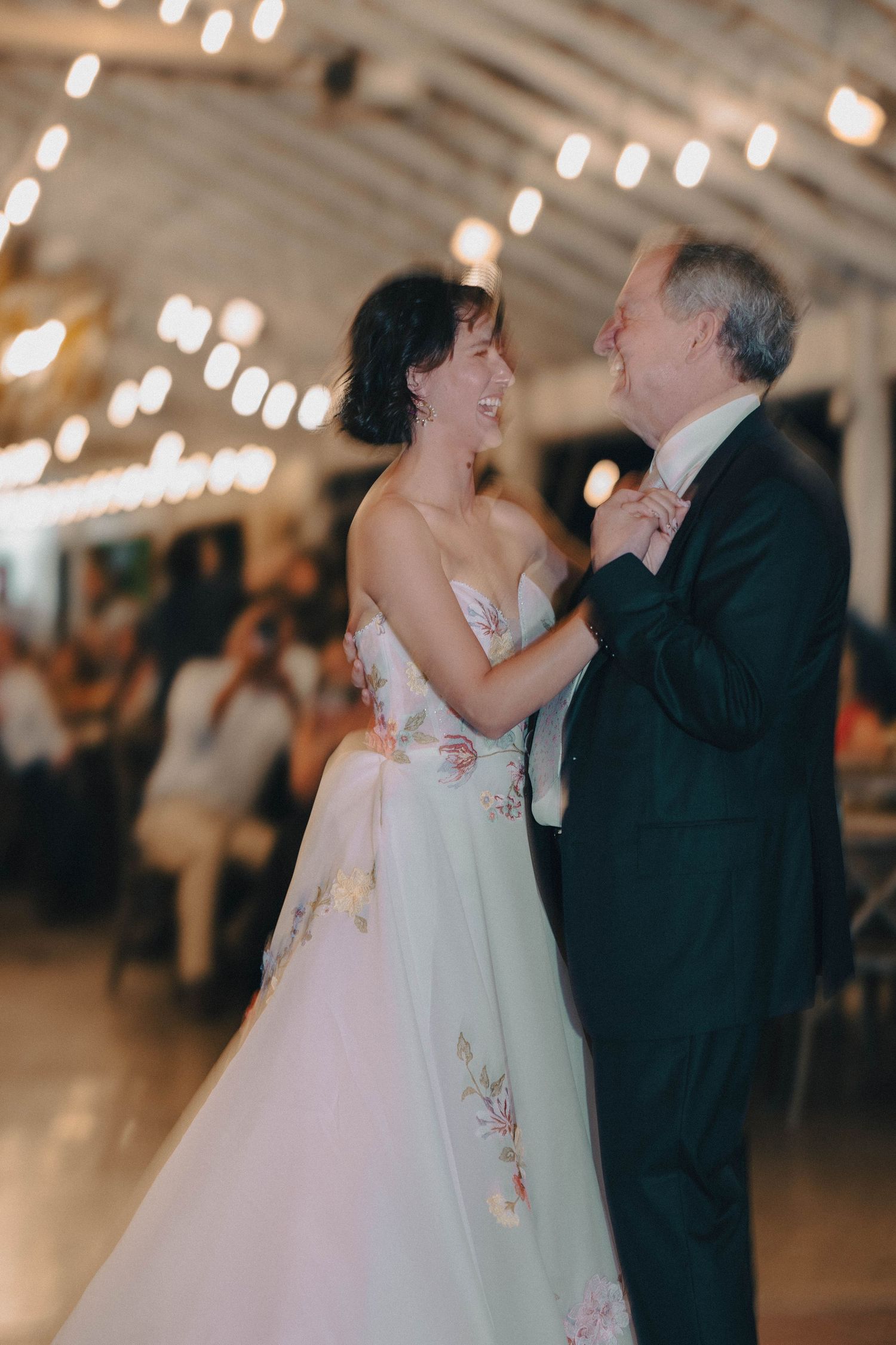 Bride in floral gown laughing while dancing with older man in dark suit at wedding reception.