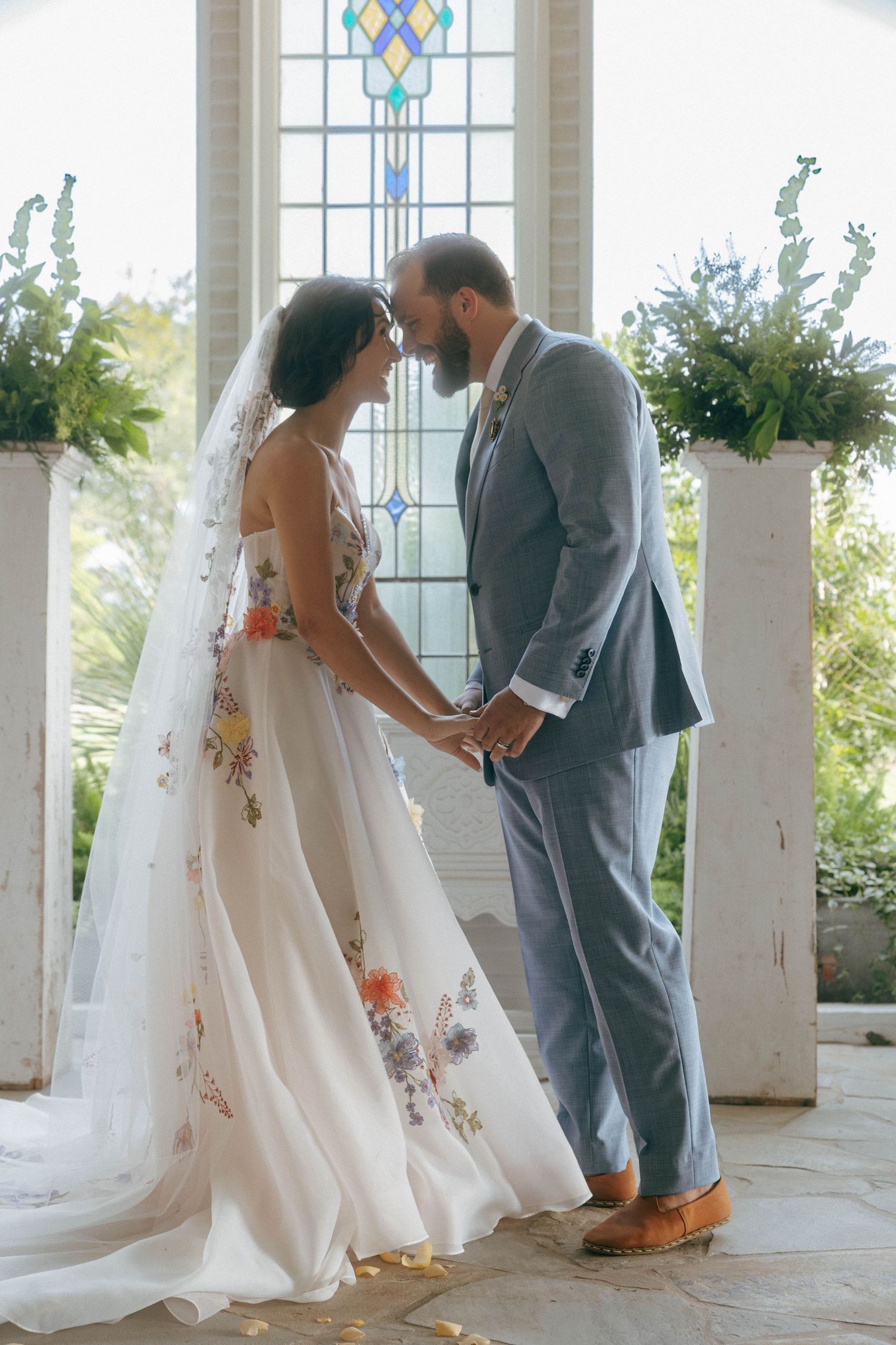 Bride and groom hold hands in a chapel with stained glass windows behind them.