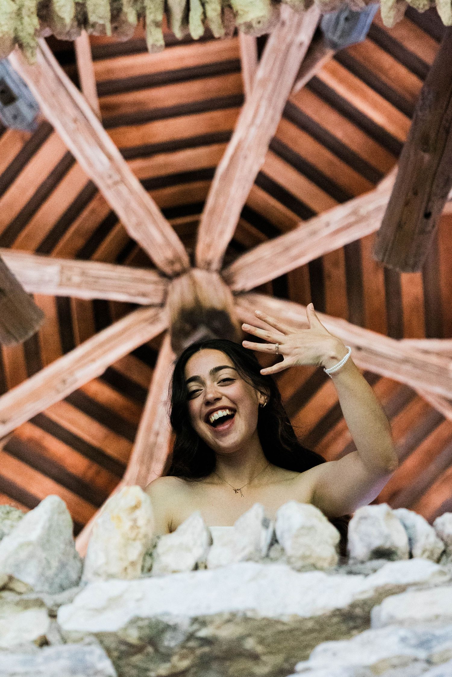Joyful bride laughing and showing engagement ring under a decorative wooden ceiling structure.