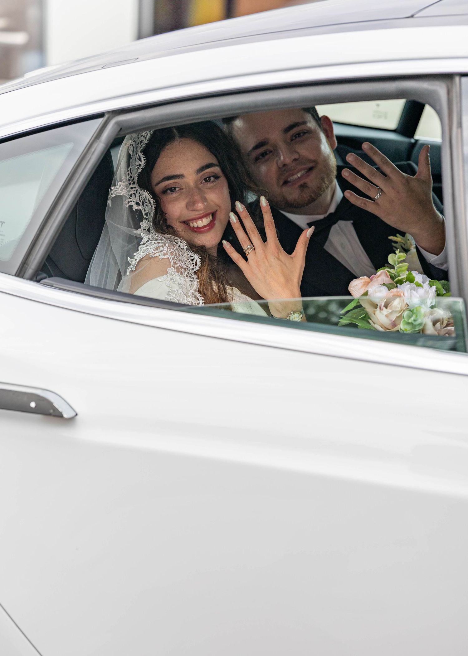 Smiling bride and groom showing wedding rings through car window with bridal bouquet visible.