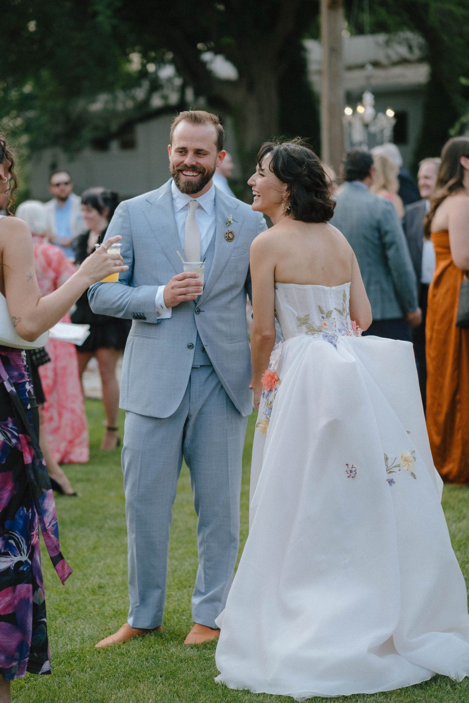 Groom in light blue suit laughs with bride in floral embroidered strapless gown at outdoor wedding reception.