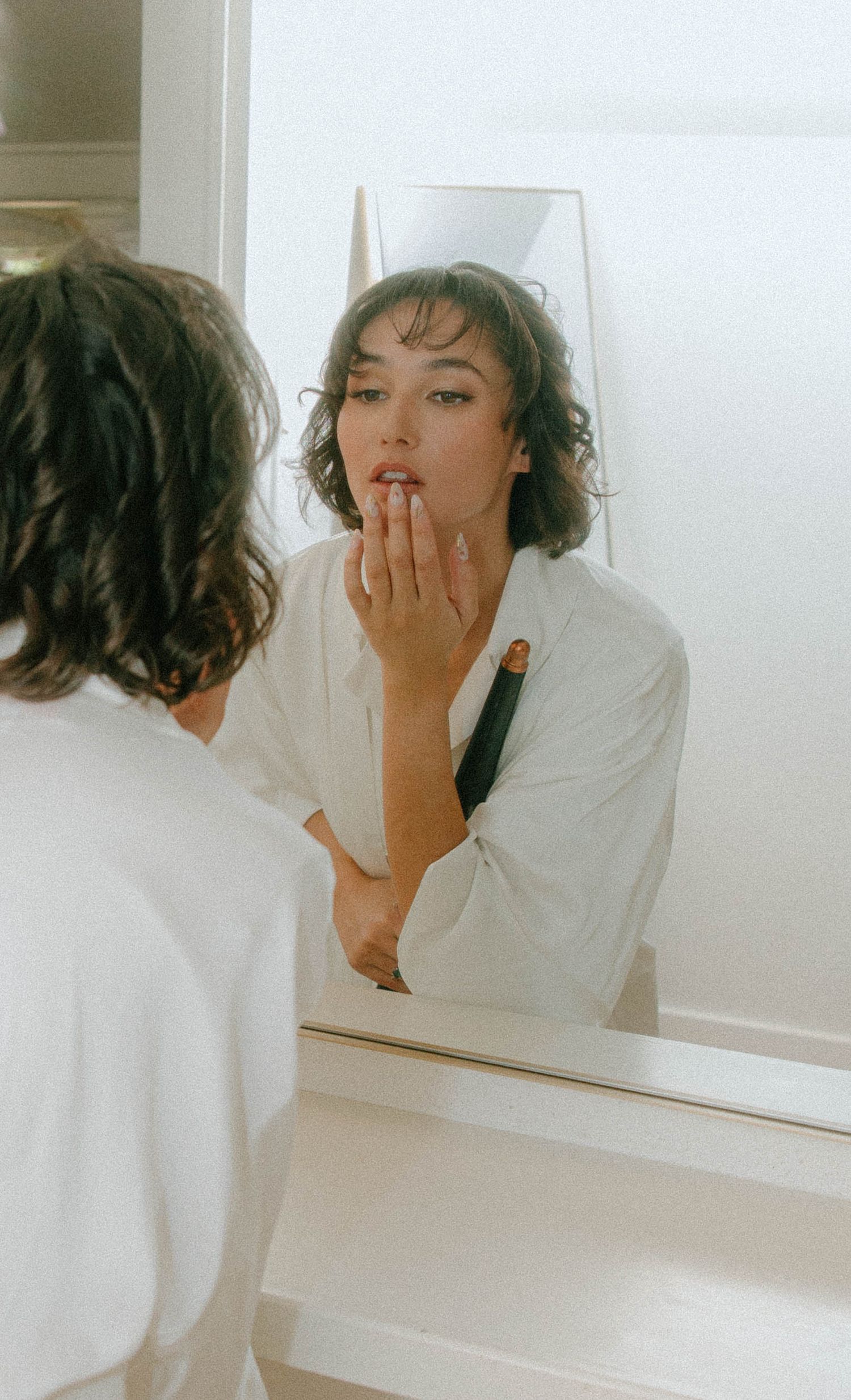 Woman with curly hair applying lipstick while looking into a mirror in natural light.
