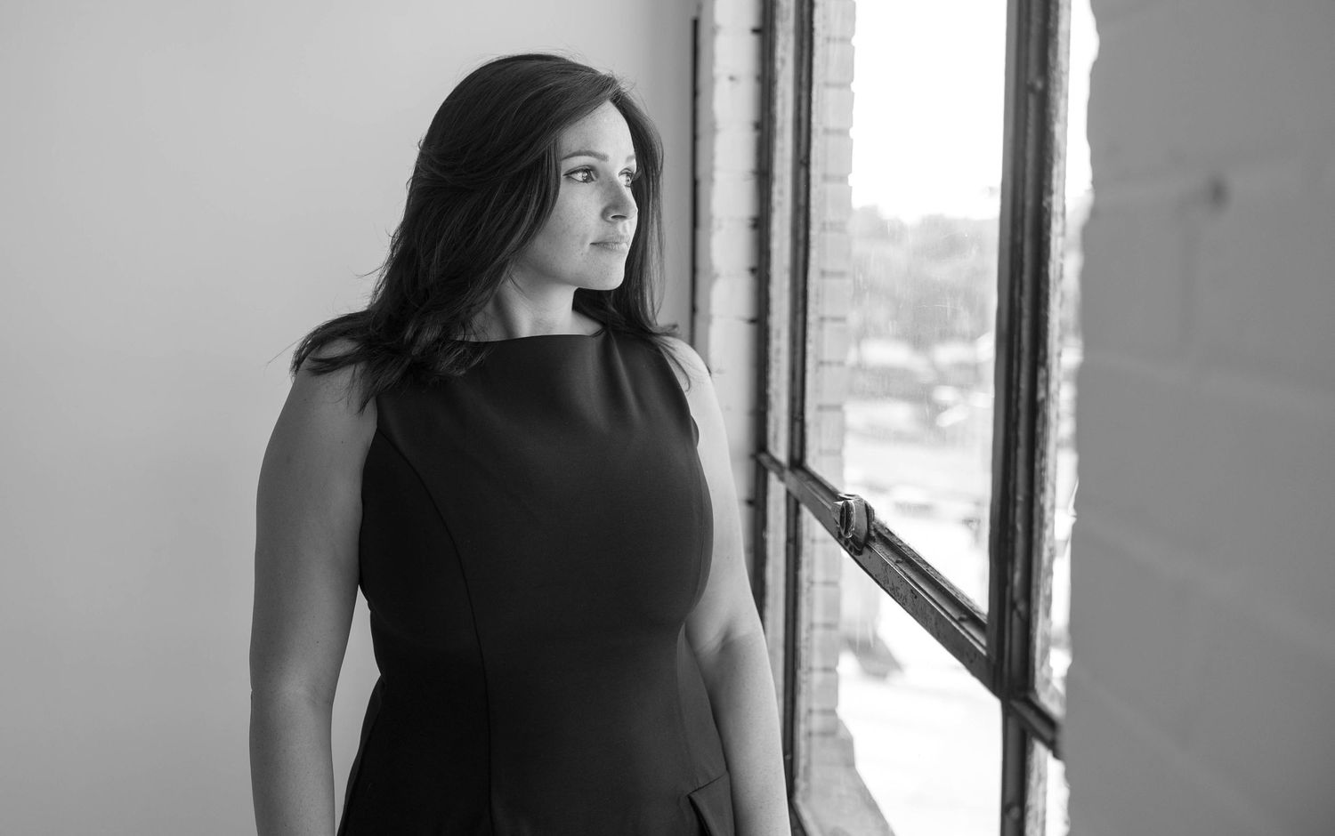 Elegant woman in black dress gazing thoughtfully out a window in black and white photo.