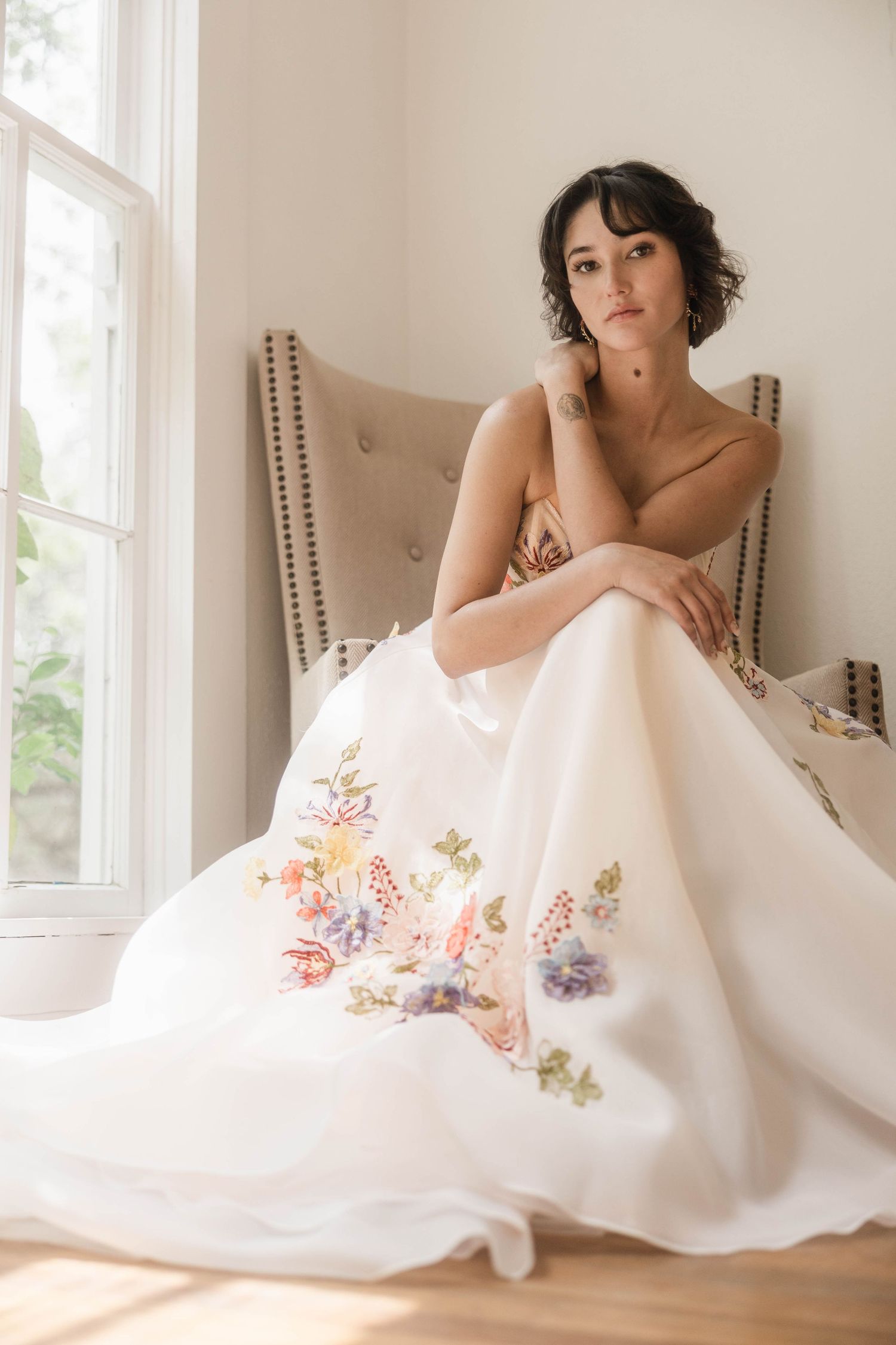 Bride in floral embroidered ball gown sits gracefully by window in elegant chair.