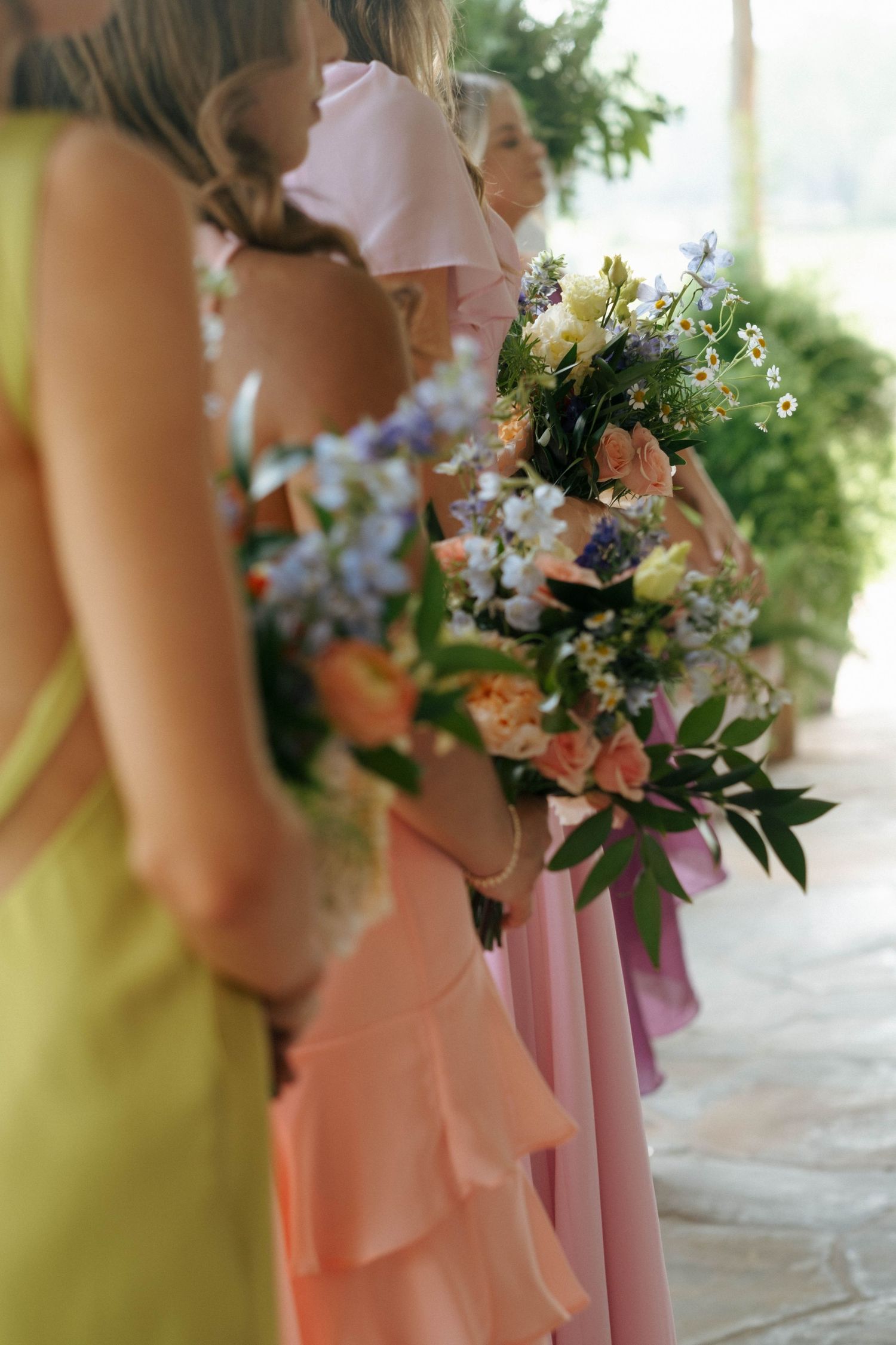 Bridesmaids in colorful mismatched dresses holding floral bouquets at a wedding ceremony.