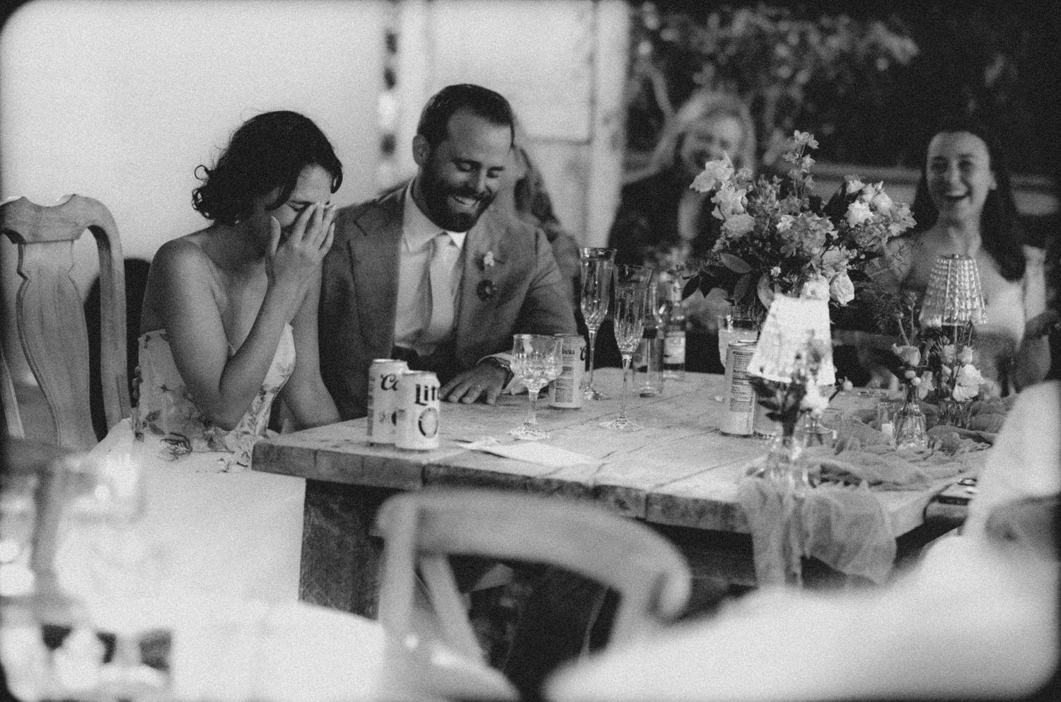 Bride and groom laugh together at their wedding reception table with floral centerpieces in black and white photo.