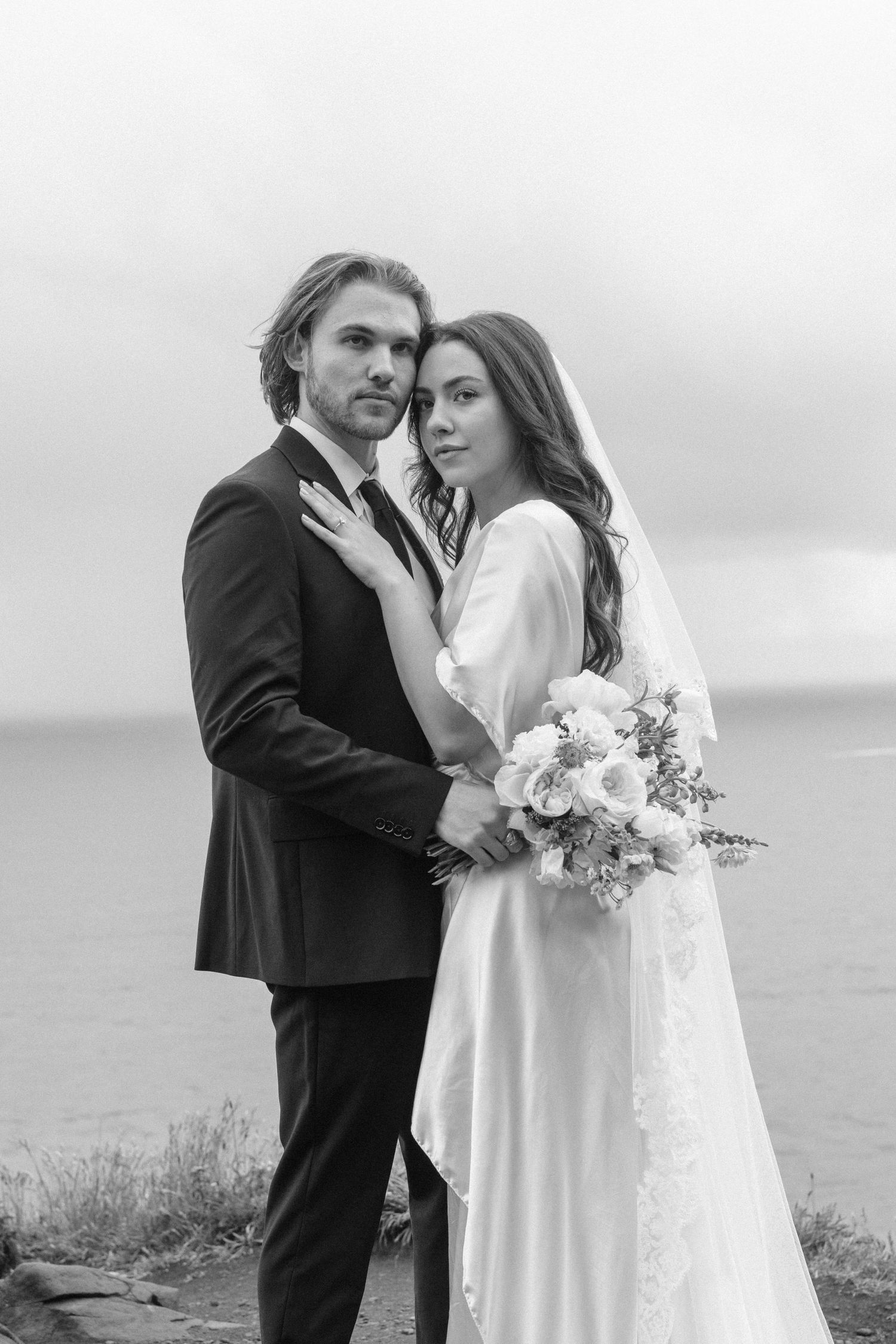 Black and white portrait of bride and groom embracing by the ocean, bride holding bouquet in satin gown with veil.