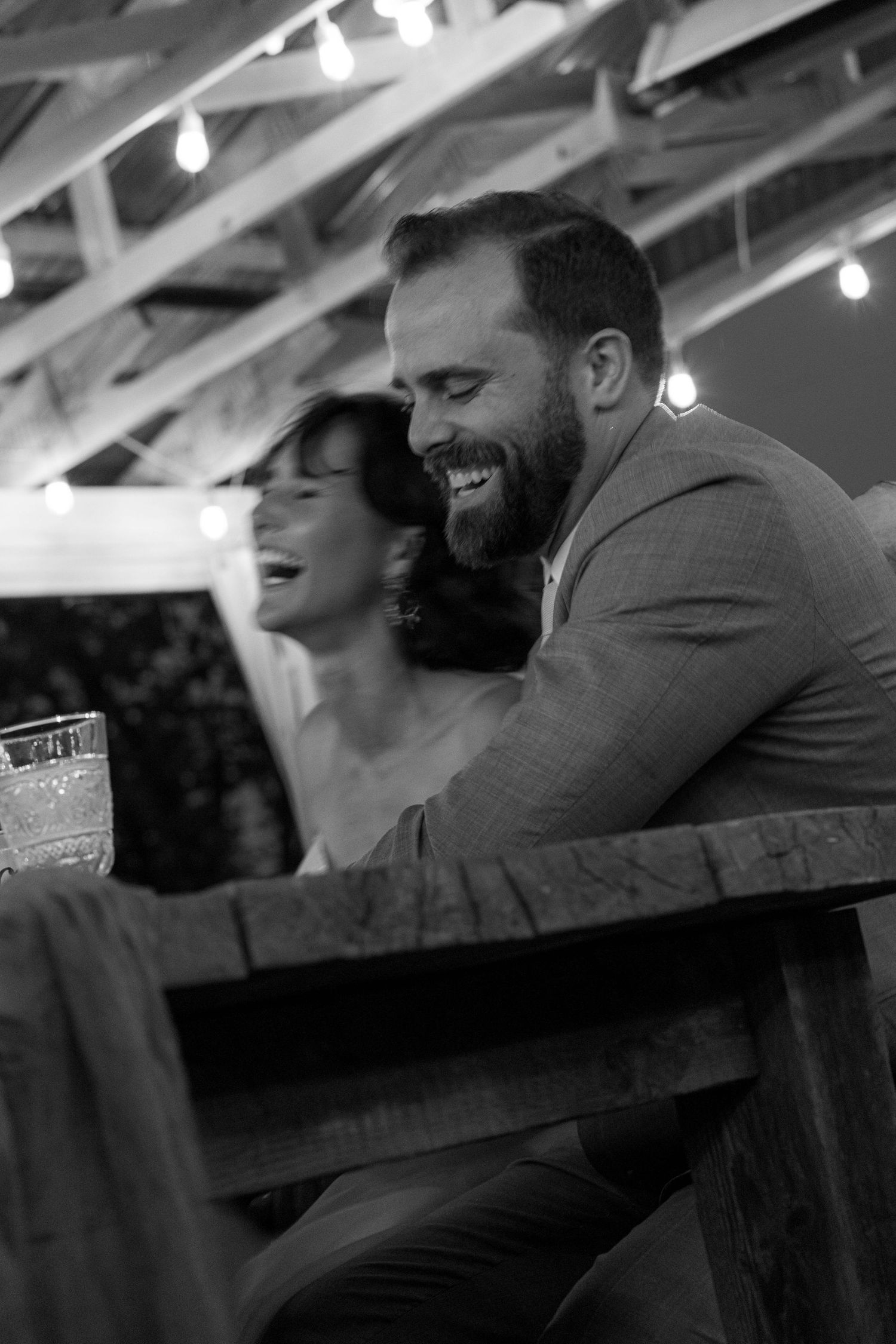 Bearded groom in a gray suit laughs joyfully alongside a woman at a rustic outdoor wedding reception.