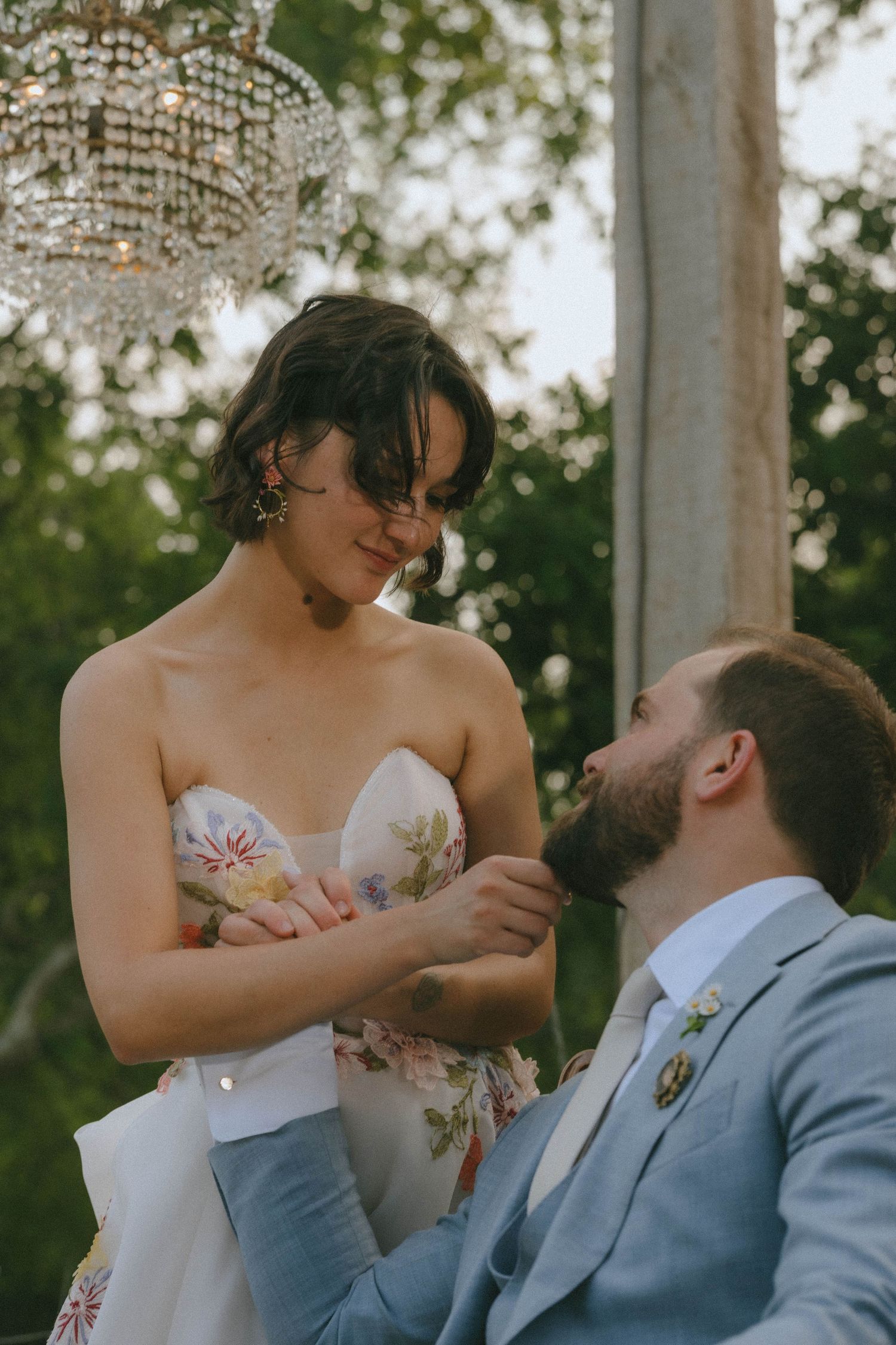 Bride in floral strapless gown tenderly touches grooms face under crystal chandelier outdoors.