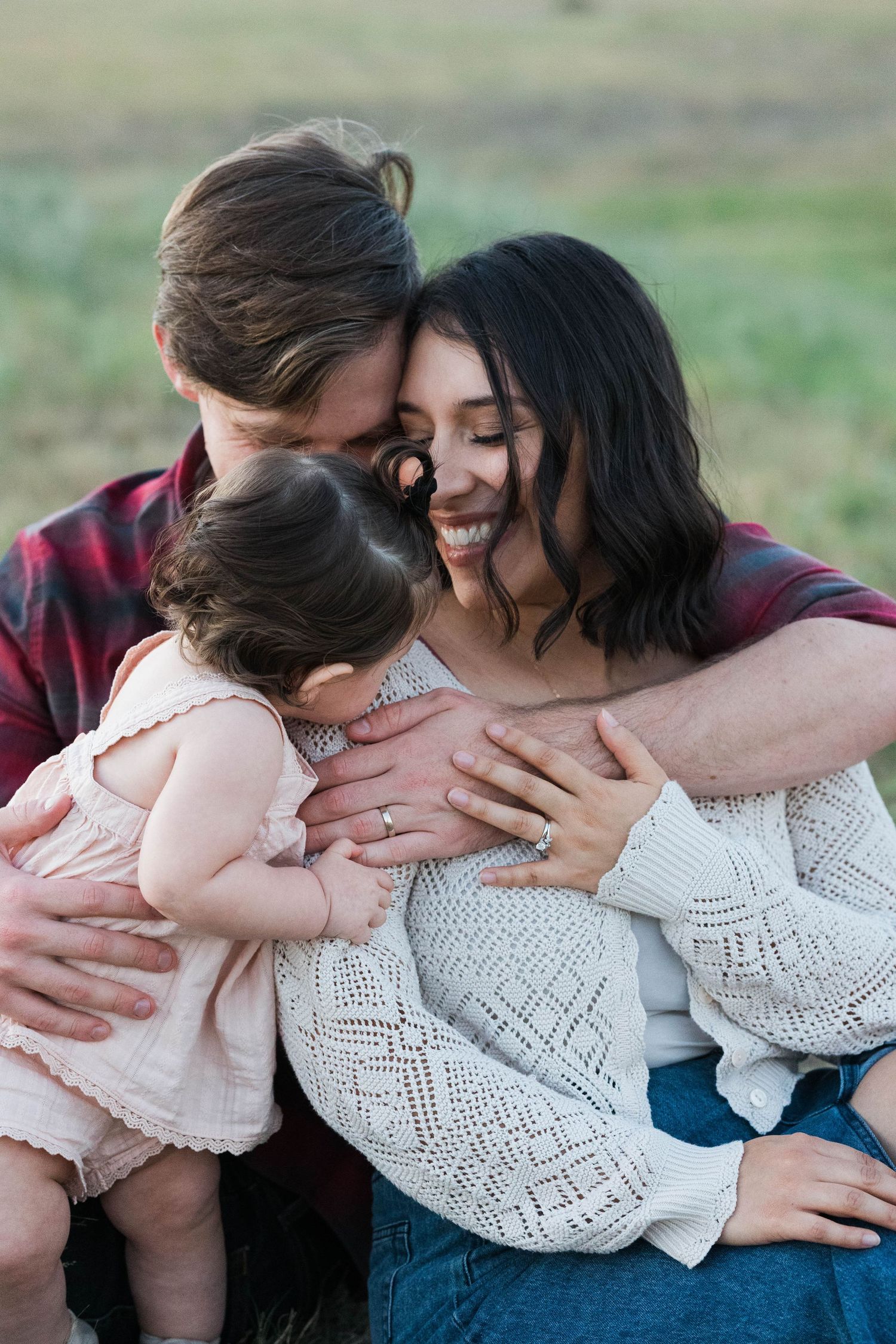 A happy young family shares a warm embrace outdoors, with mother in cream sweater and father in plaid shirt.