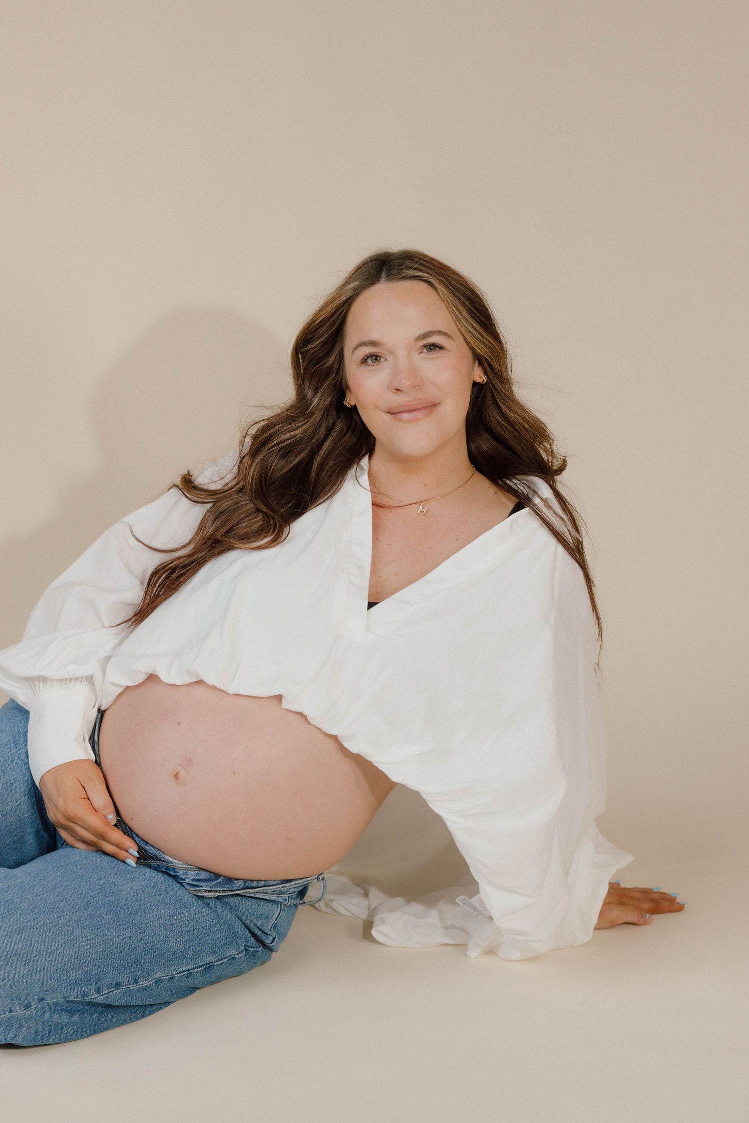 Serene pregnant woman in white blouse and jeans sitting on floor holding baby bump.