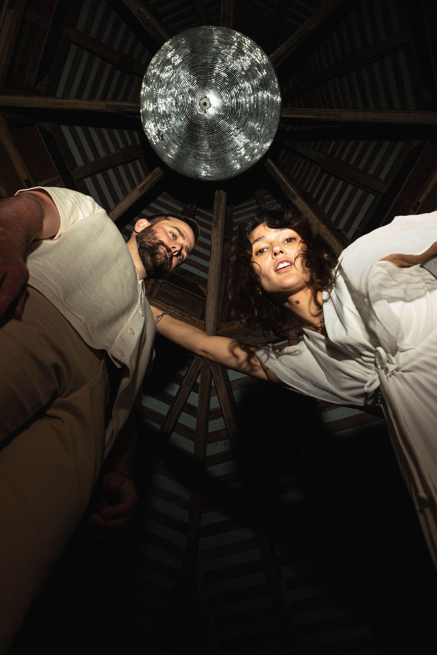Couple photographed from below inside a wooden gazebo, smiling down at camera with a disco ball overhead.