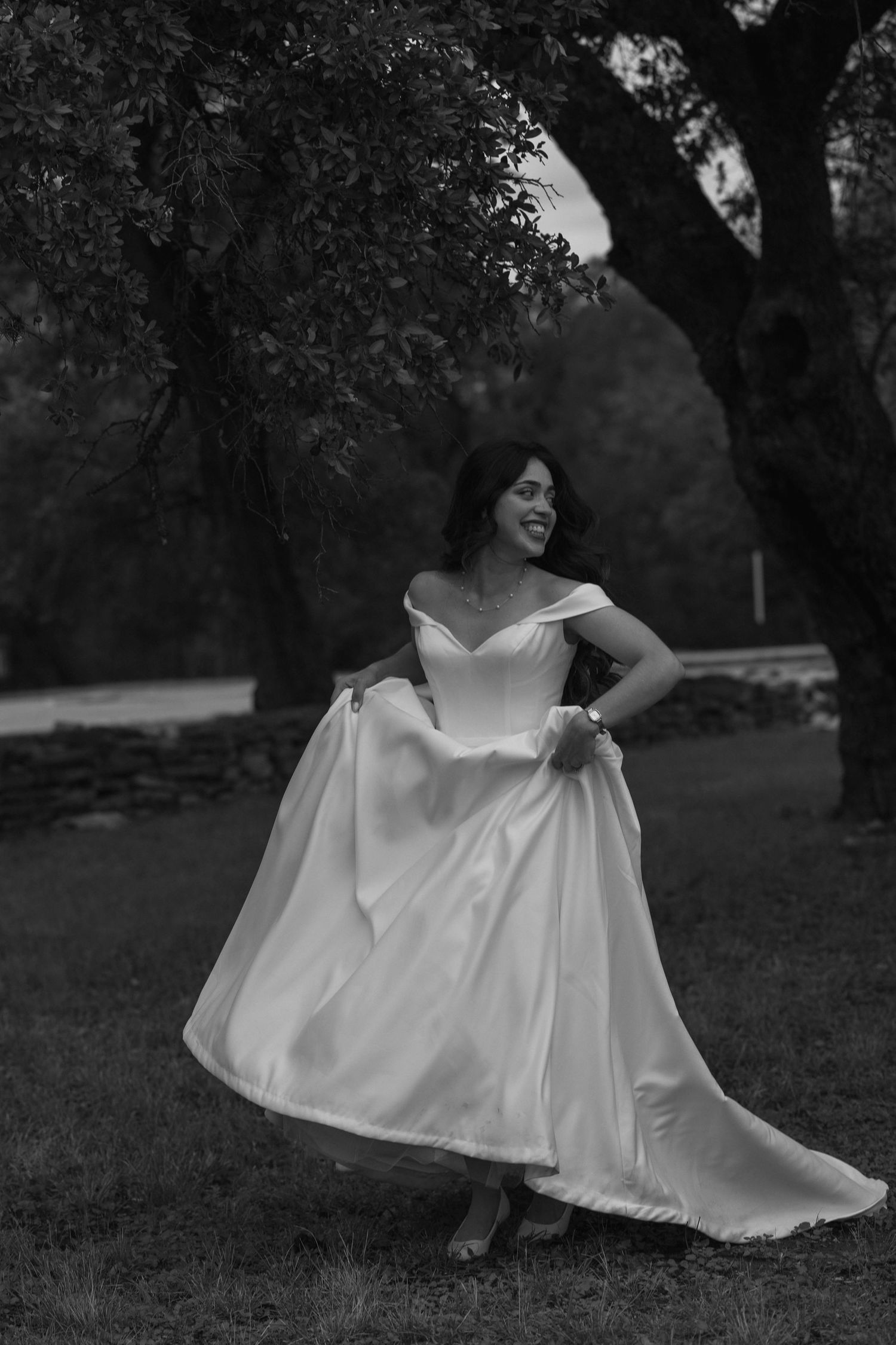 Bride in off-shoulder wedding gown twirls joyfully under trees in a black and white outdoor portrait.
