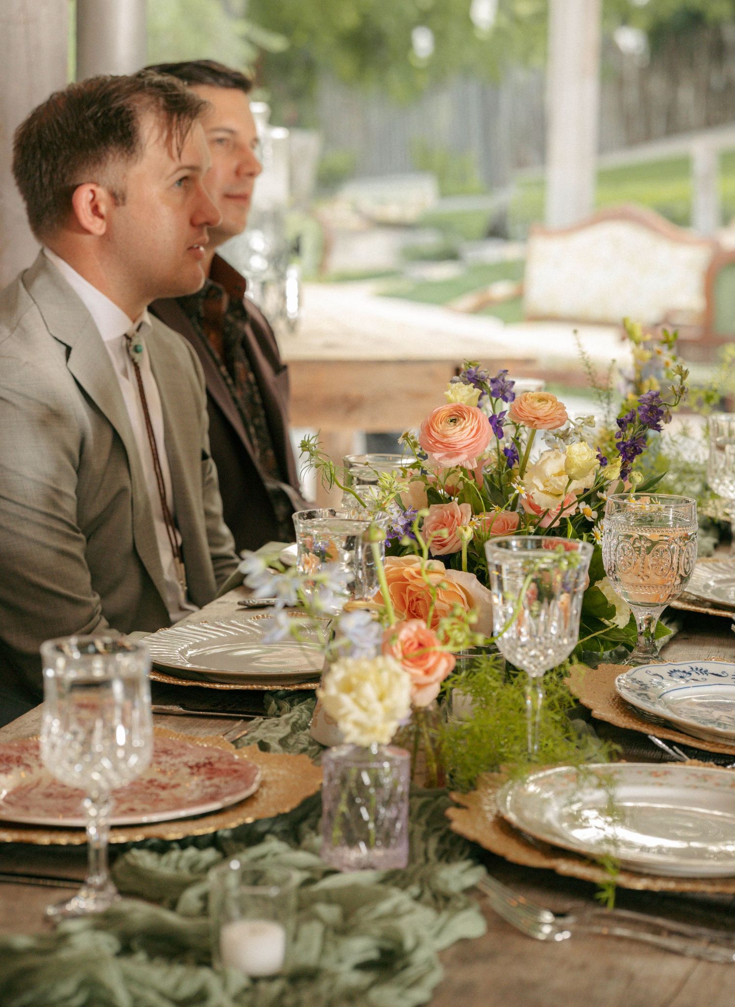 Two men in suits seated at an elegantly decorated wedding reception table with floral centerpiece.