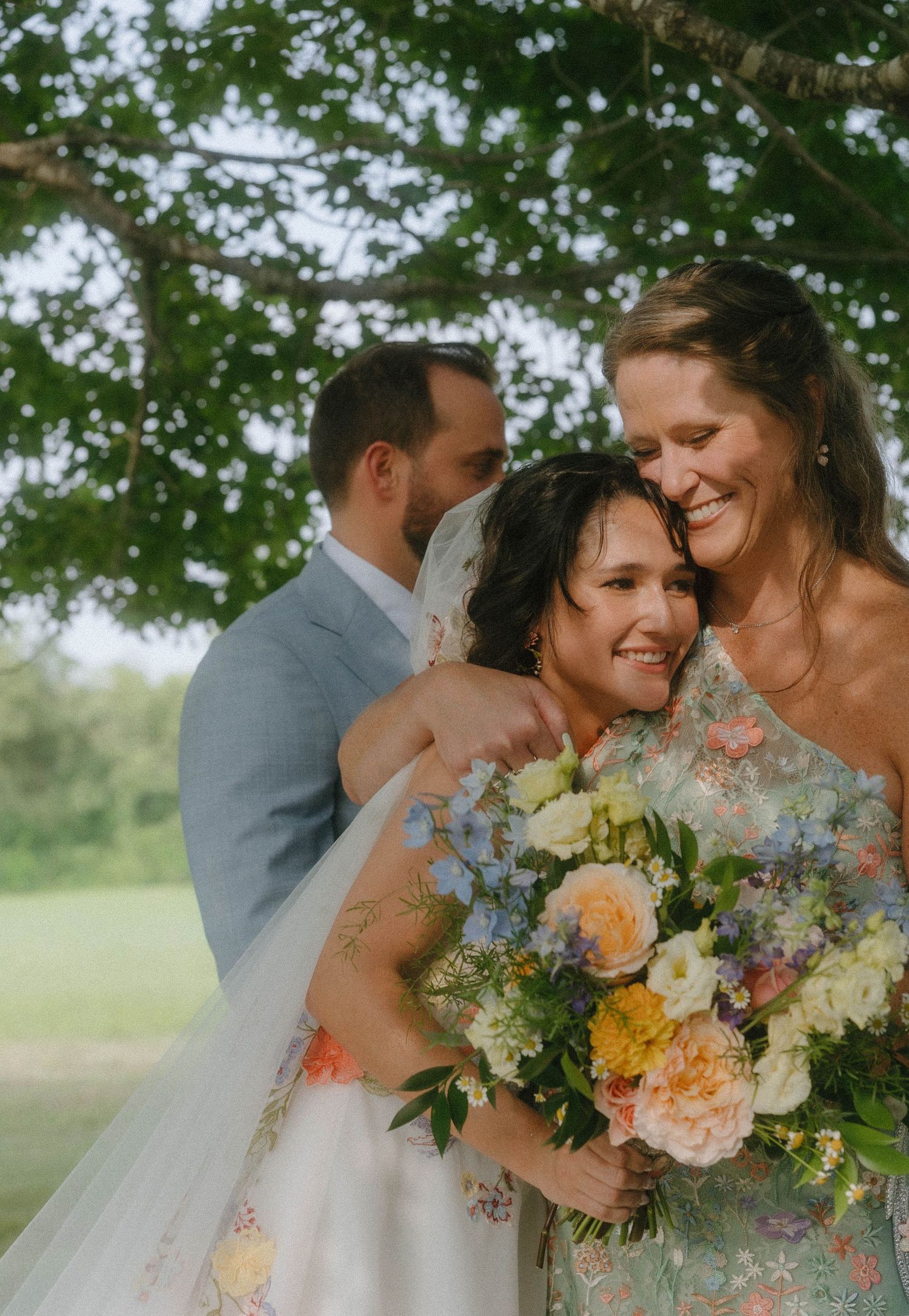 Bride in floral wedding gown embraces woman while groom stands behind under green tree.