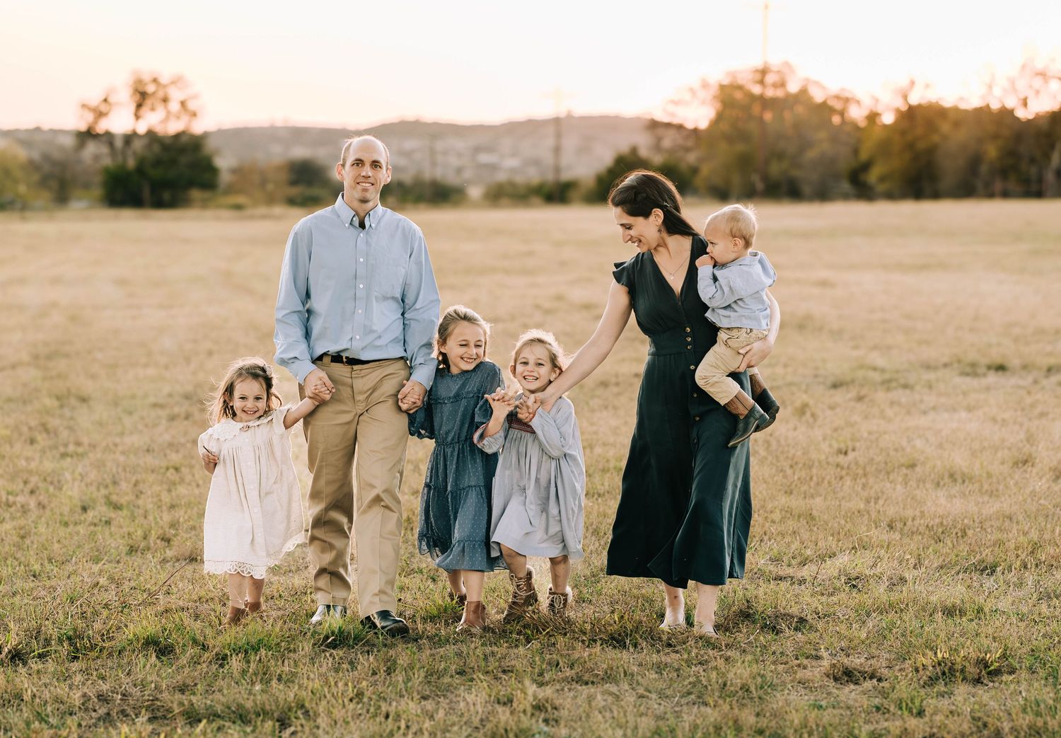 Happy family of six walking through a sunny open field at golden hour, mother carrying baby while children hold hands.