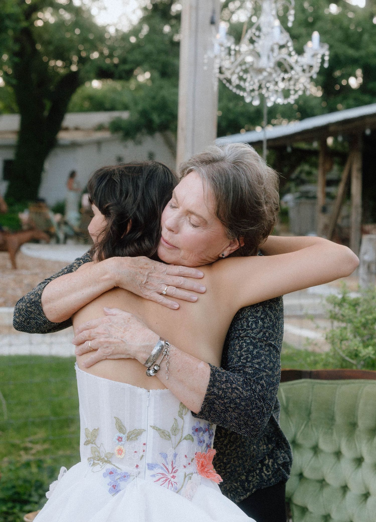 Bride in floral strapless gown sharing emotional hug with older woman at outdoor wedding.