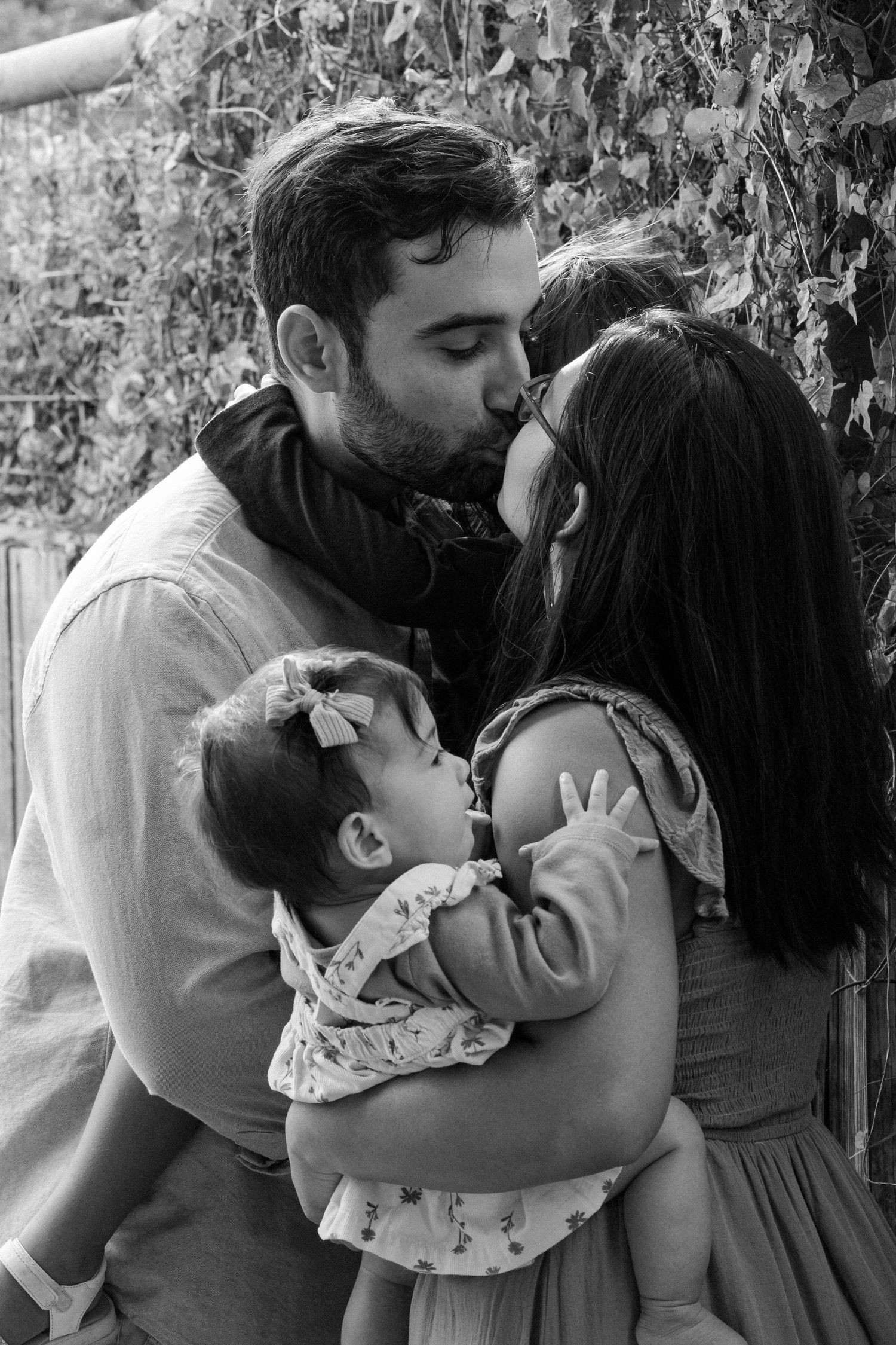 Black and white photo of a couple kissing while holding their baby and young daughter outdoors.