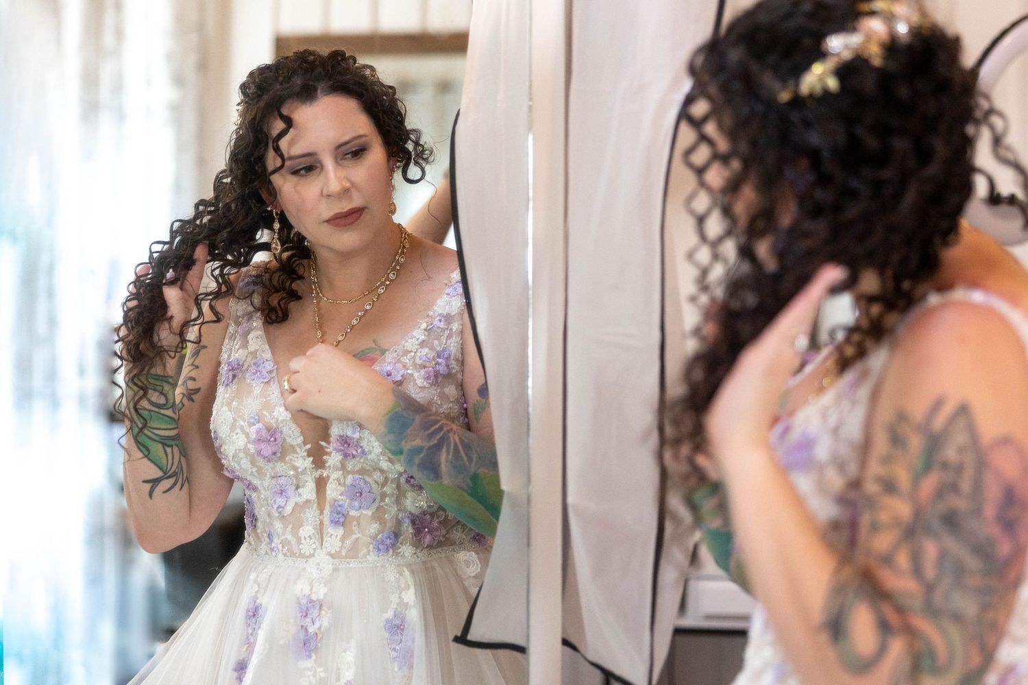 Tattooed bride with curly hair adjusts her floral wedding gown while looking in a mirror before the ceremony.