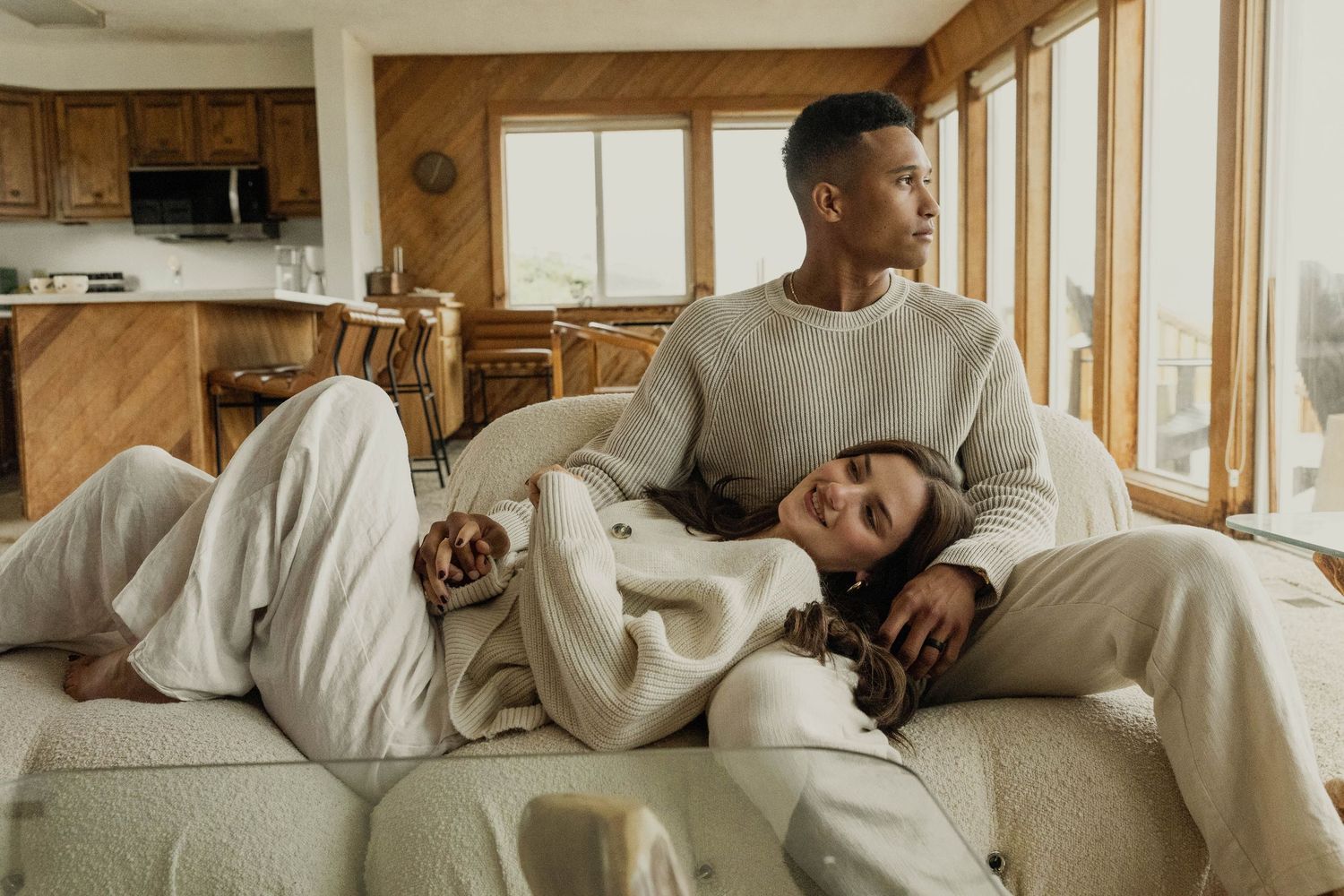 A relaxed couple lounges on a cream sofa in a cozy beach house living room, gazing into the distance.