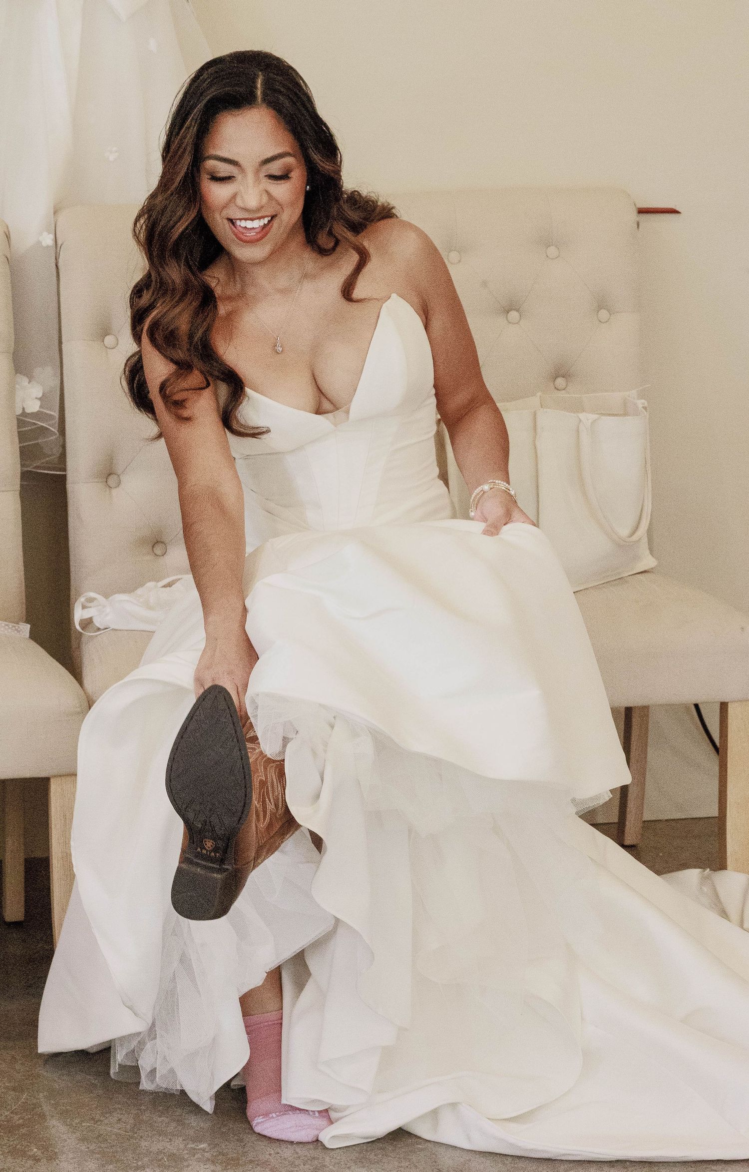 Joyful bride in strapless white ball gown laughing while putting on her wedding shoes.