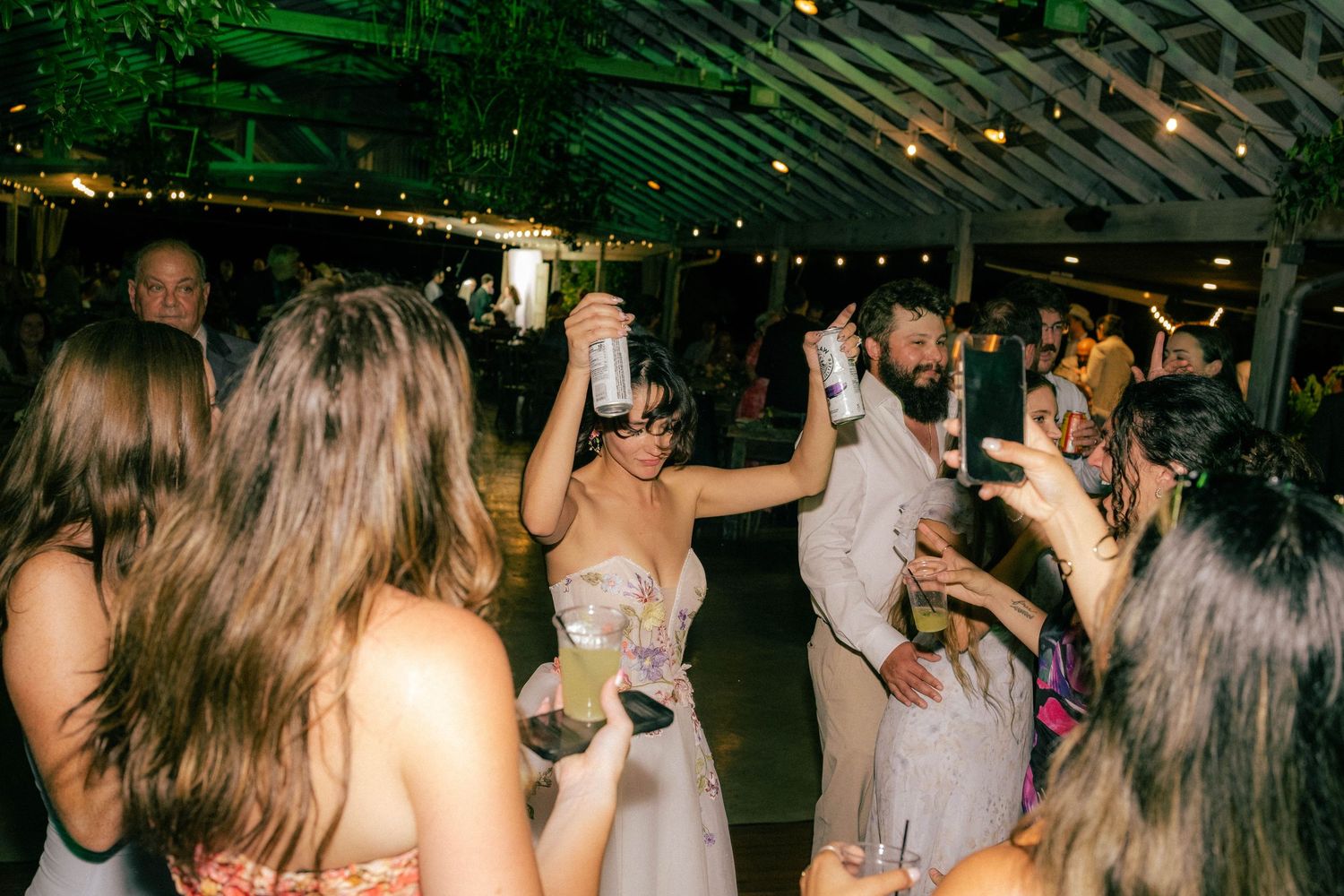 Bride and groom celebrate with raised drinks surrounded by dancing guests under string lights at wedding reception.