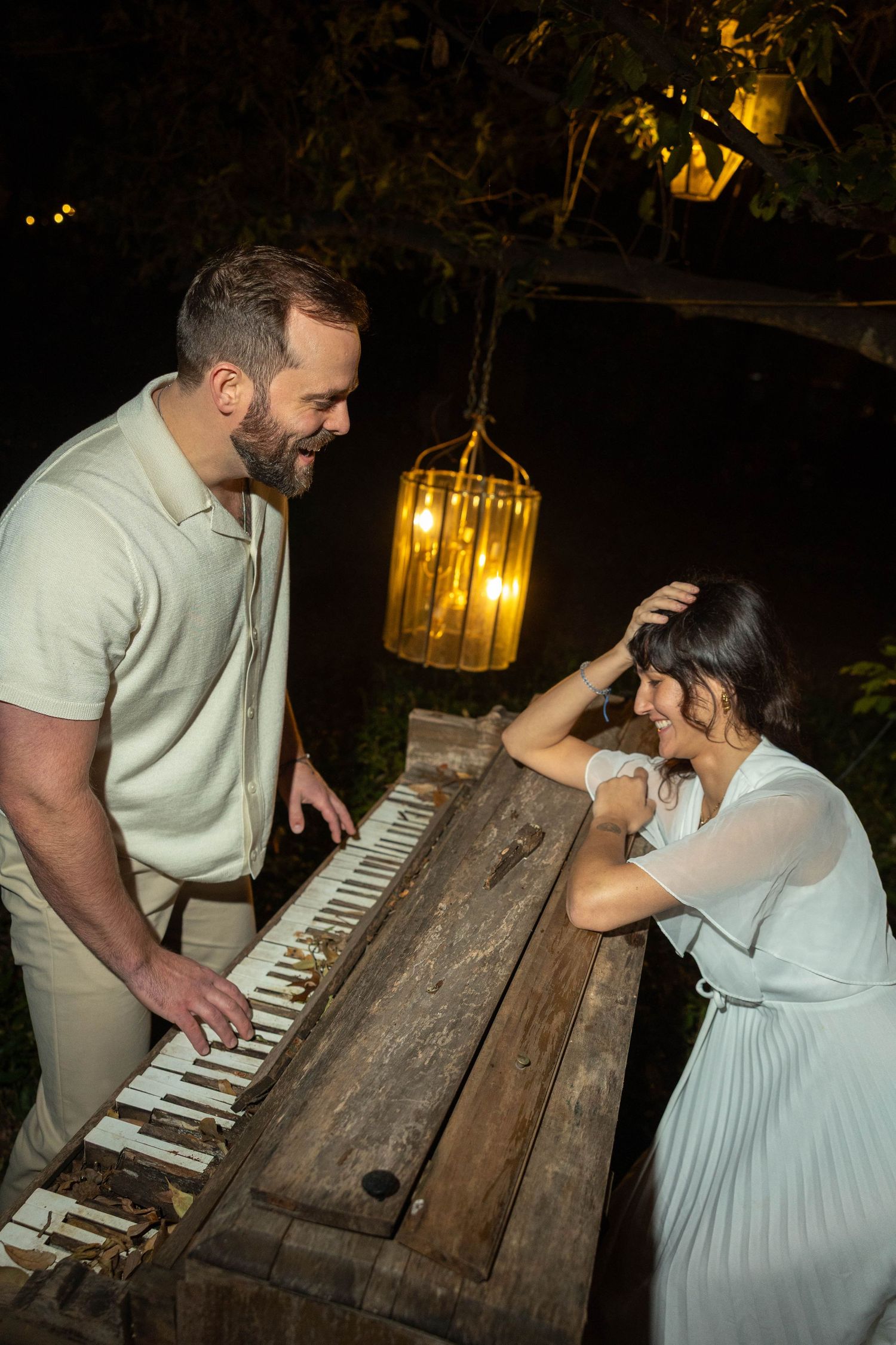 Couple laughing together at a weathered outdoor piano under golden lantern light at night in a garden setting.