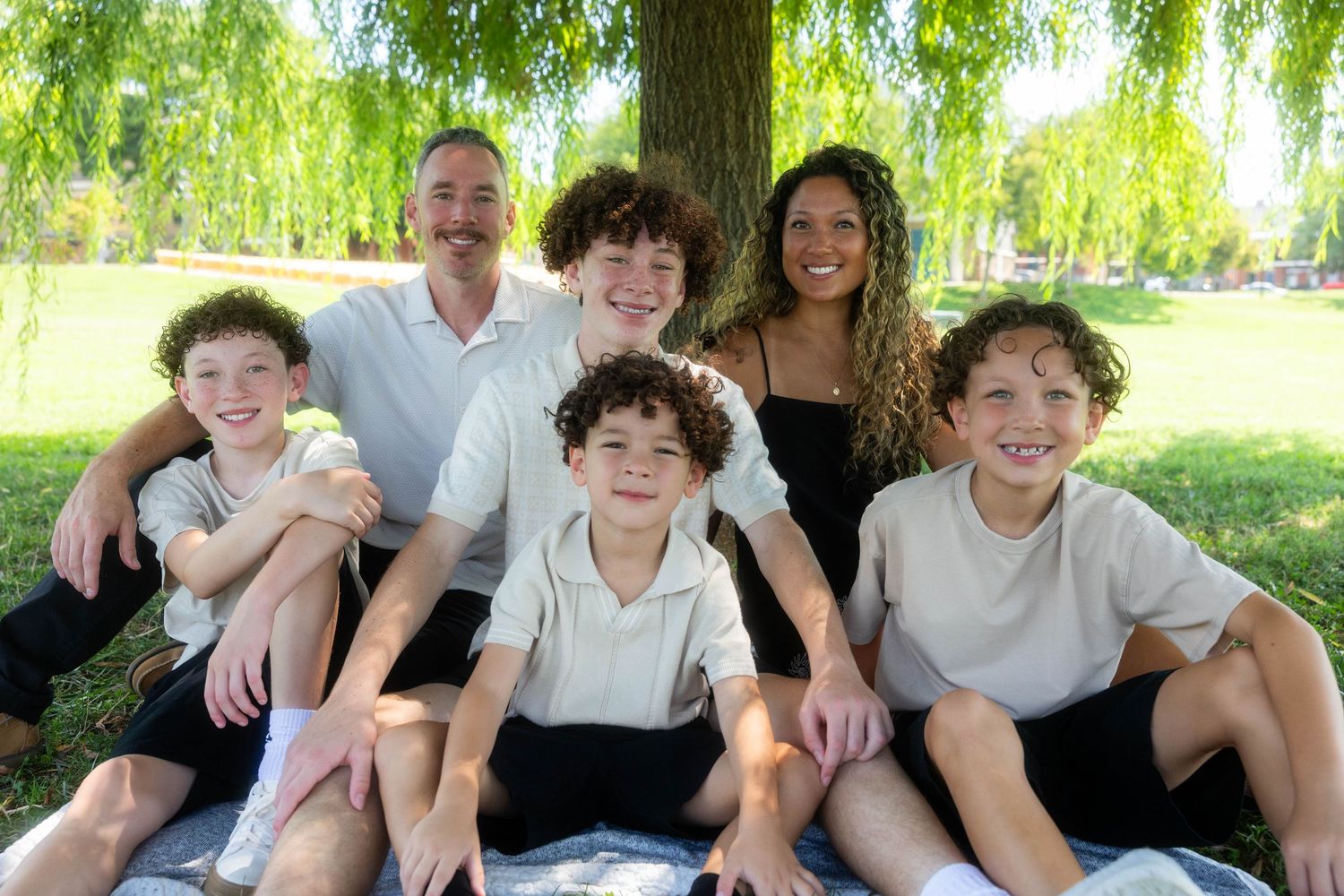 Family of six smiling outdoors under a willow tree on a sunny day, parents with four curly-haired boys.