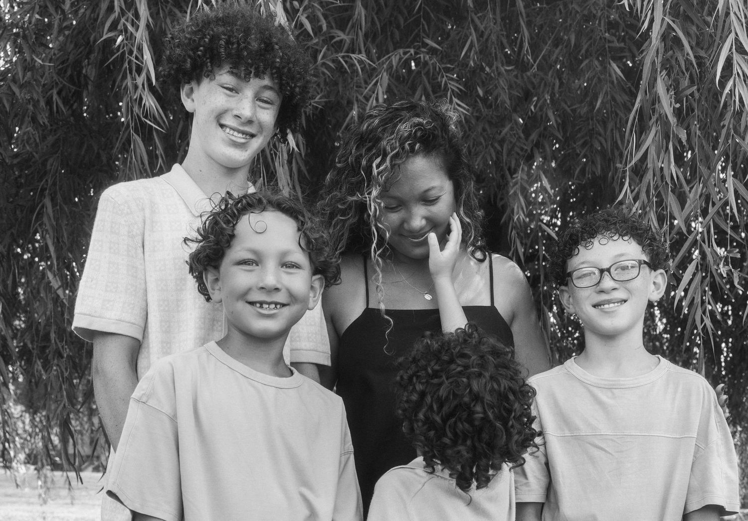 Black and white photo of five smiling children posing together outdoors under a weeping willow tree.