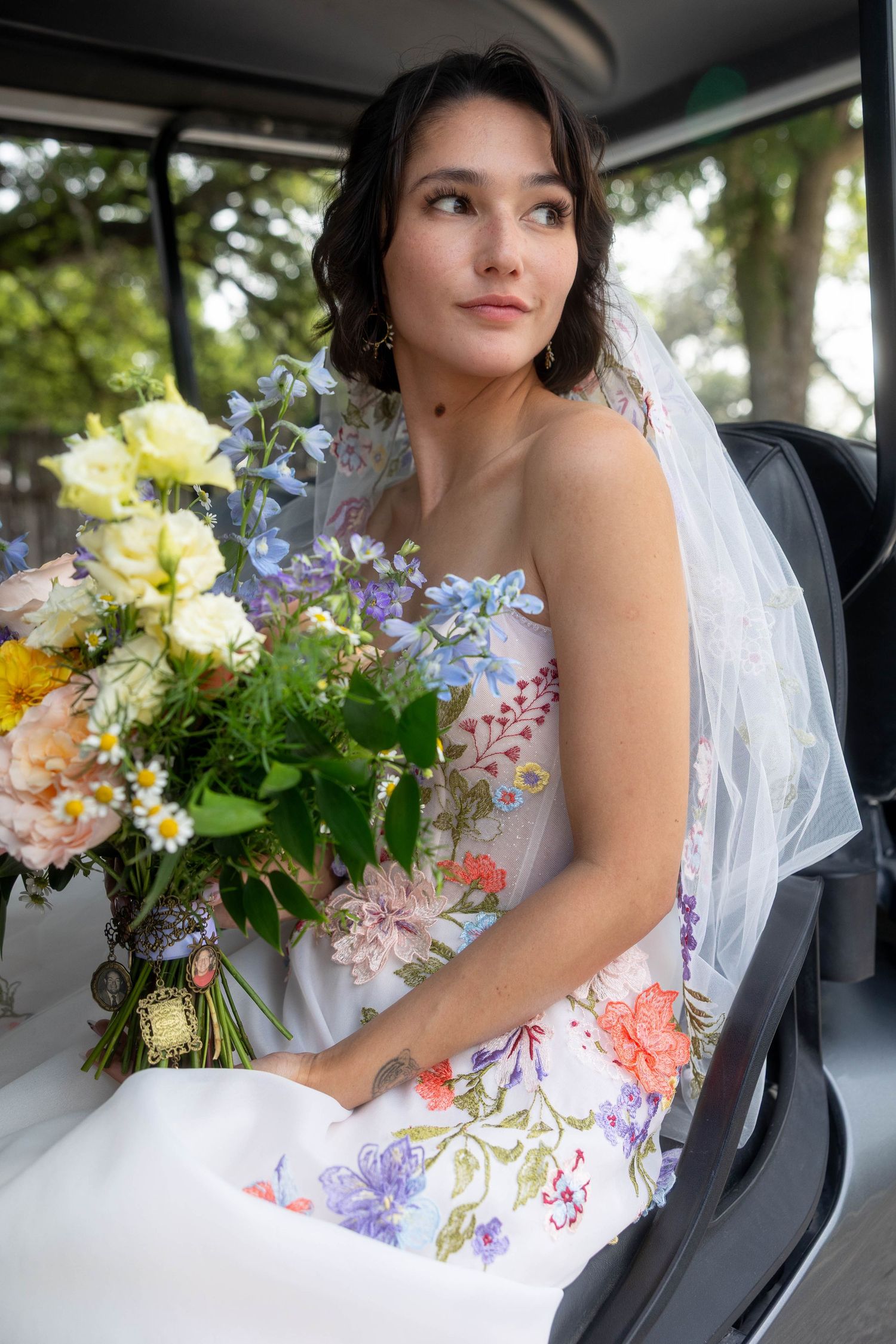 Bride in floral wedding gown holding colorful bouquet while seated in vehicle with veil.