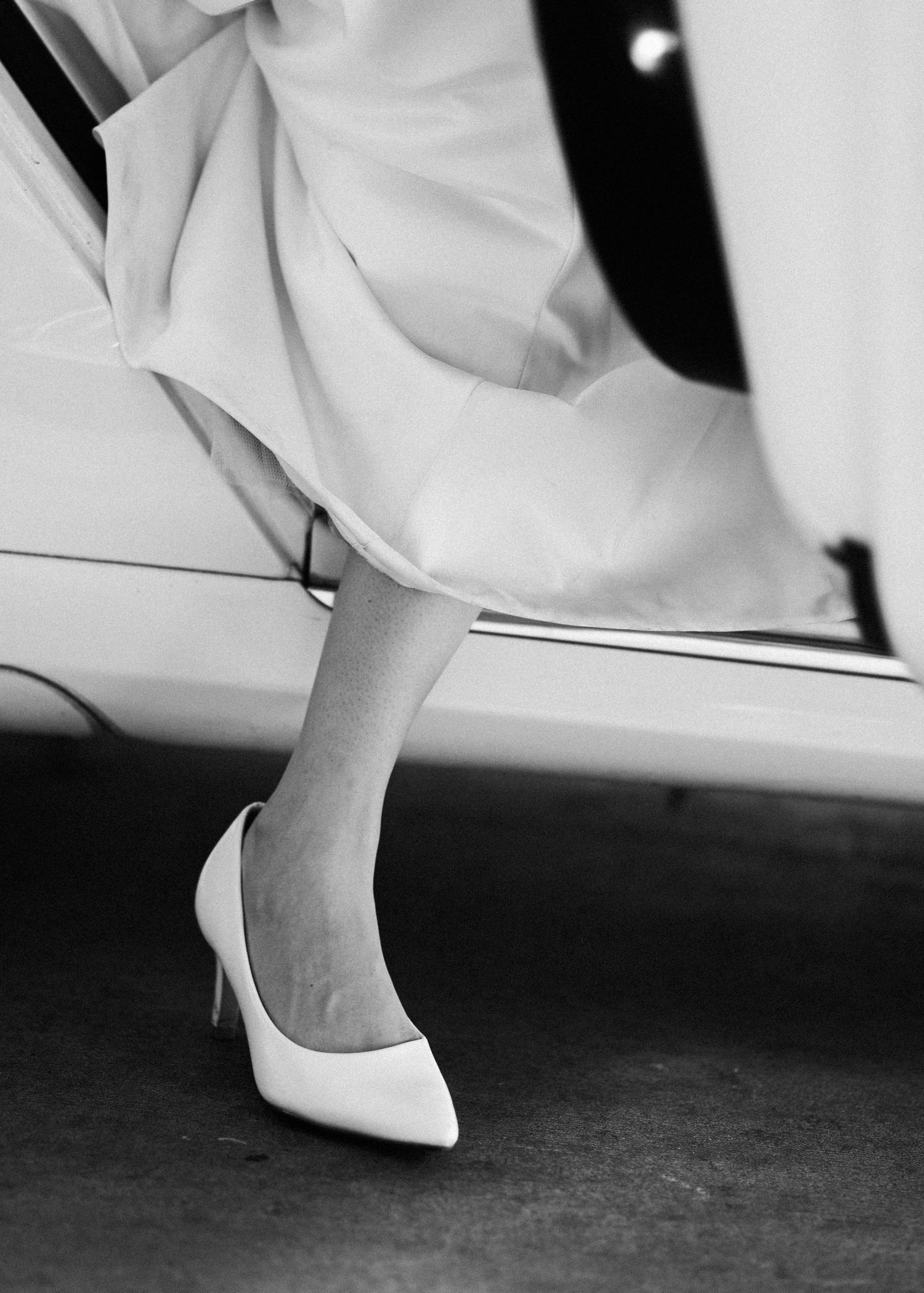 Black and white close-up of a bride stepping out of a car in elegant white pointed-toe heels.