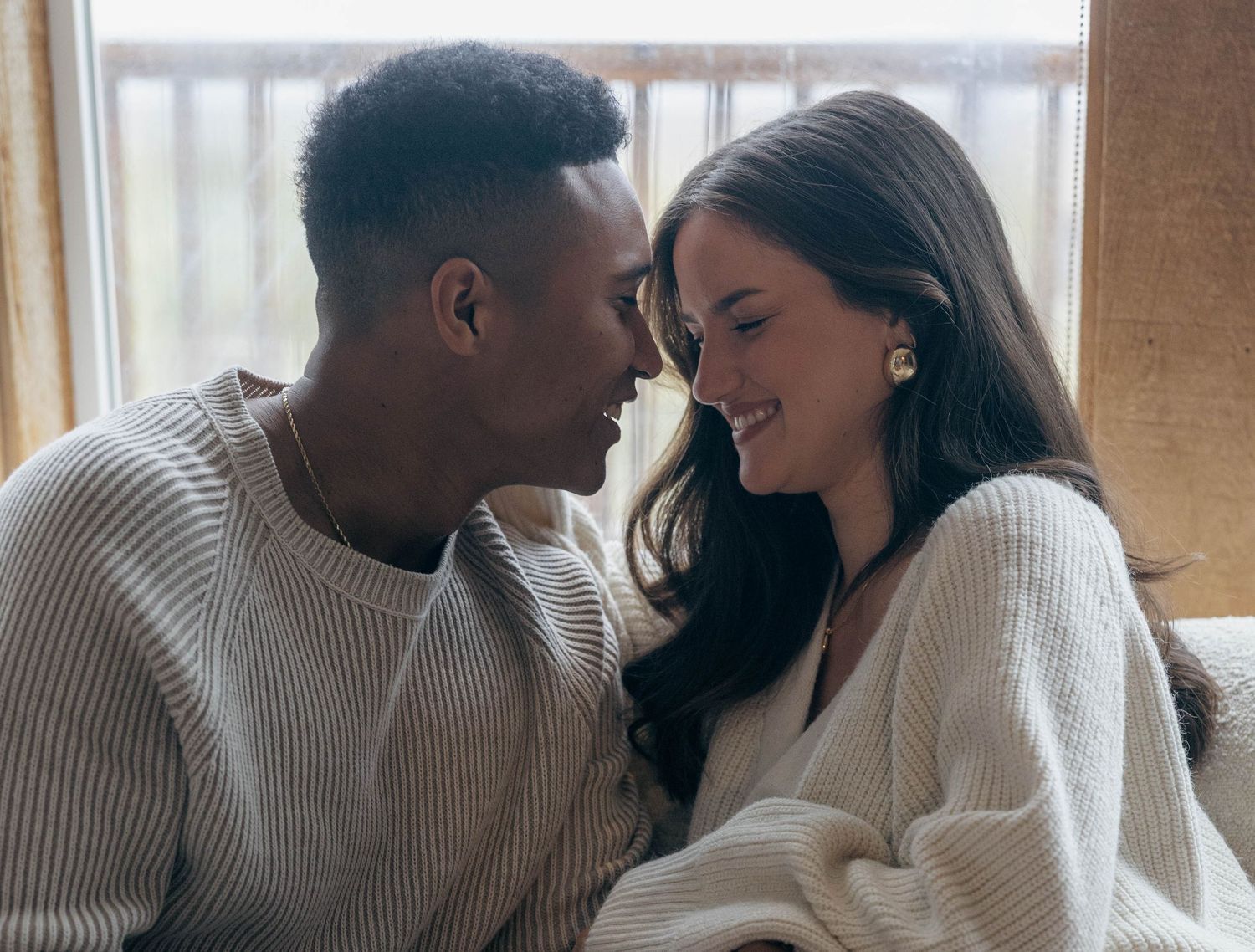A smiling couple shares a tender moment together, with the woman wearing gold earrings in a warm interior.