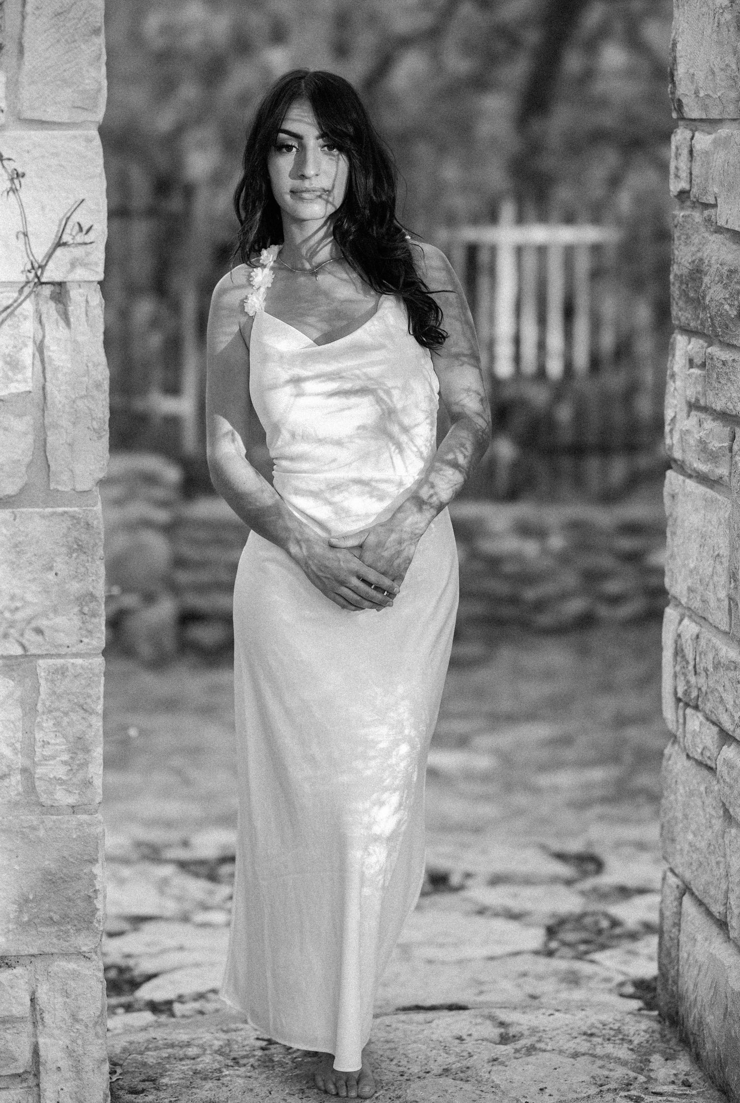Black and white photo of bride in white slip dress standing between stone columns.