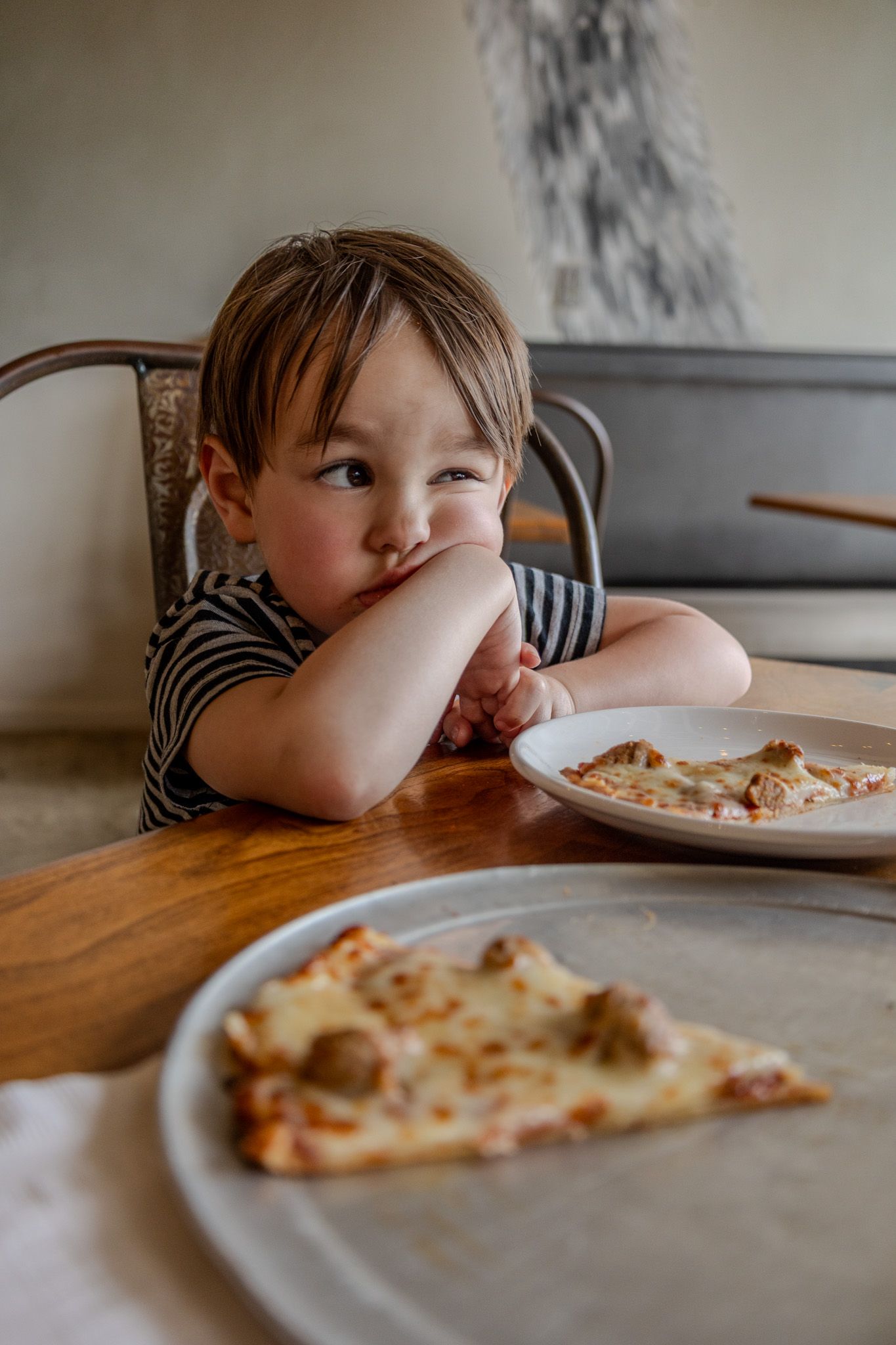 Toddler sitting at wooden table with pizza, resting chin on hands with thoughtful expression.