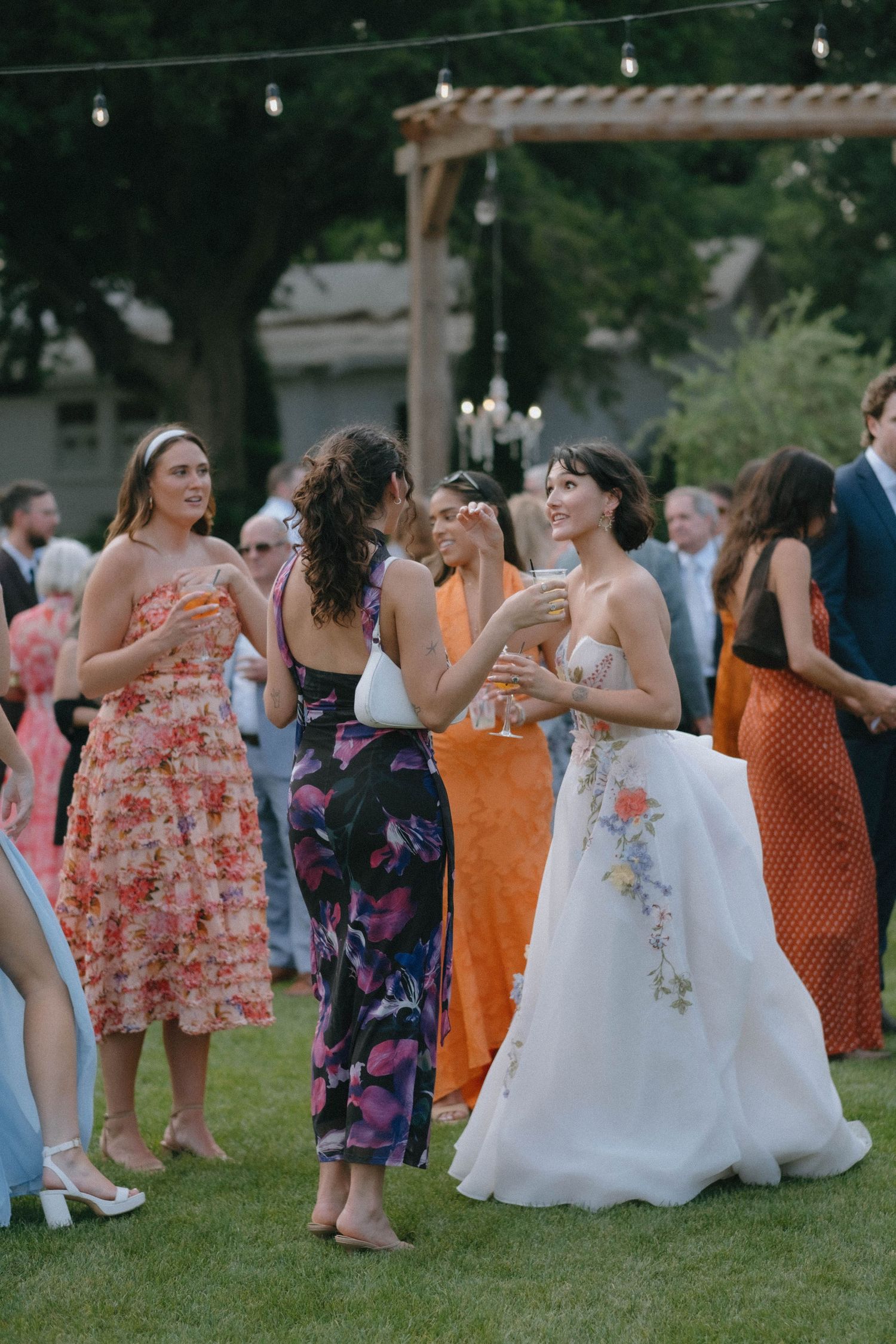 Bride in floral embroidered white gown mingles with guests in colorful dresses at an outdoor wedding reception.