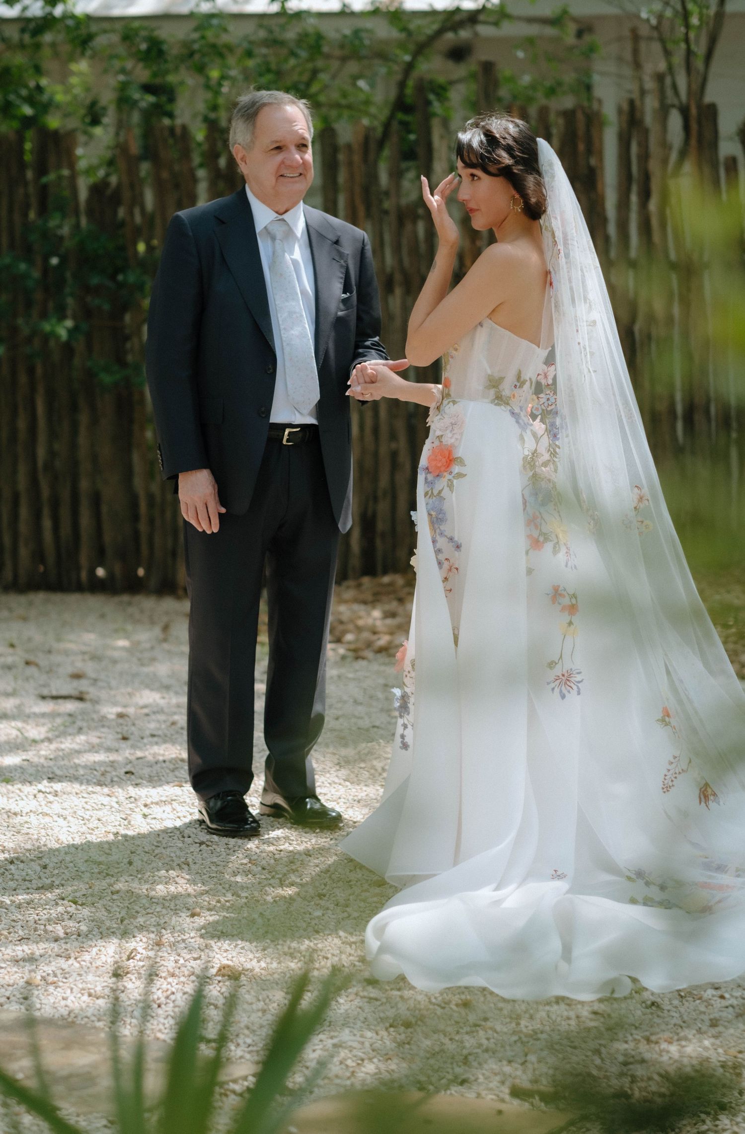 Emotional bride in a floral embroidered wedding gown and veil wiping tears while holding hands with her father.